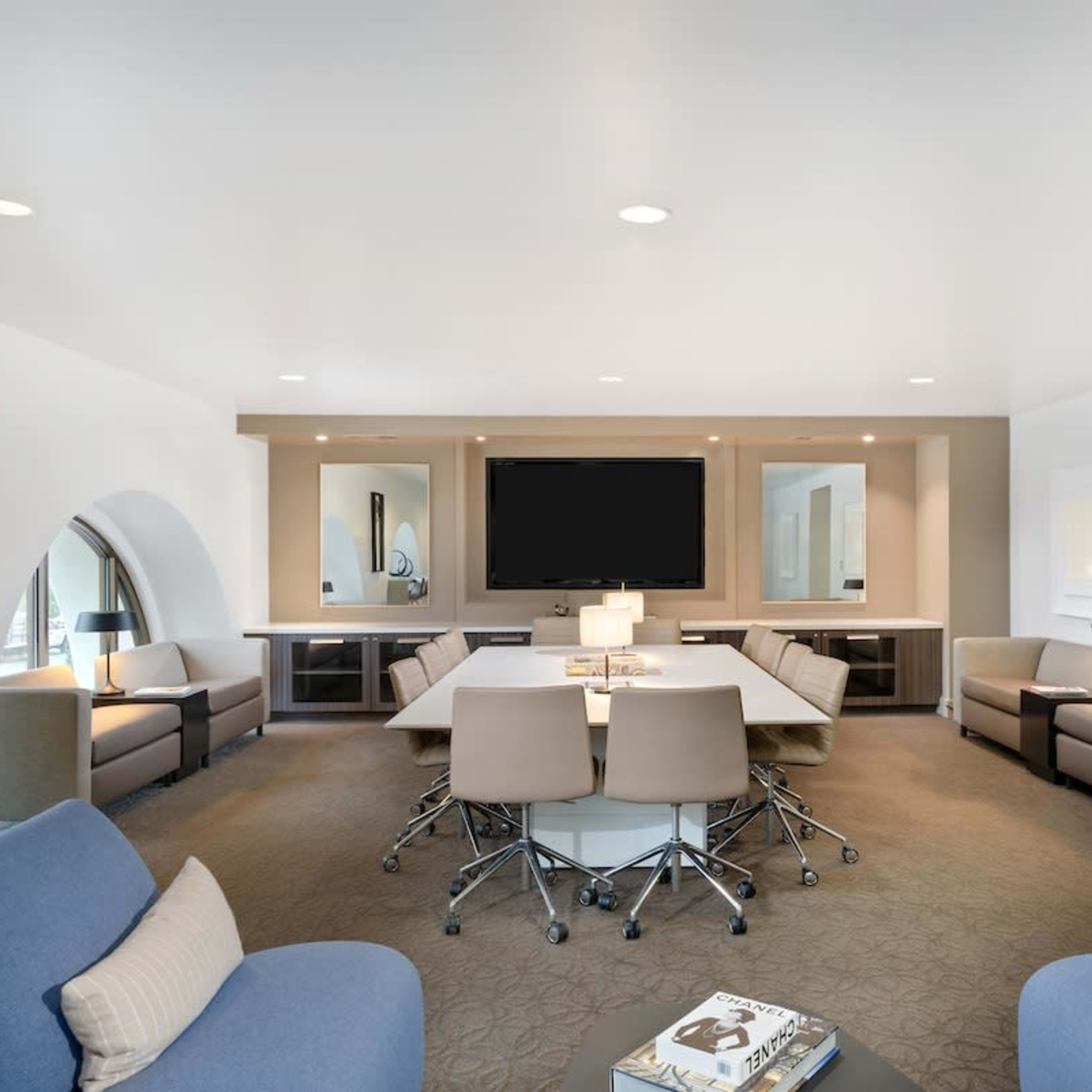A modern conference room featuring a central table surrounded by rolling chairs, with a large screen mounted on the wall and seating areas on either side.