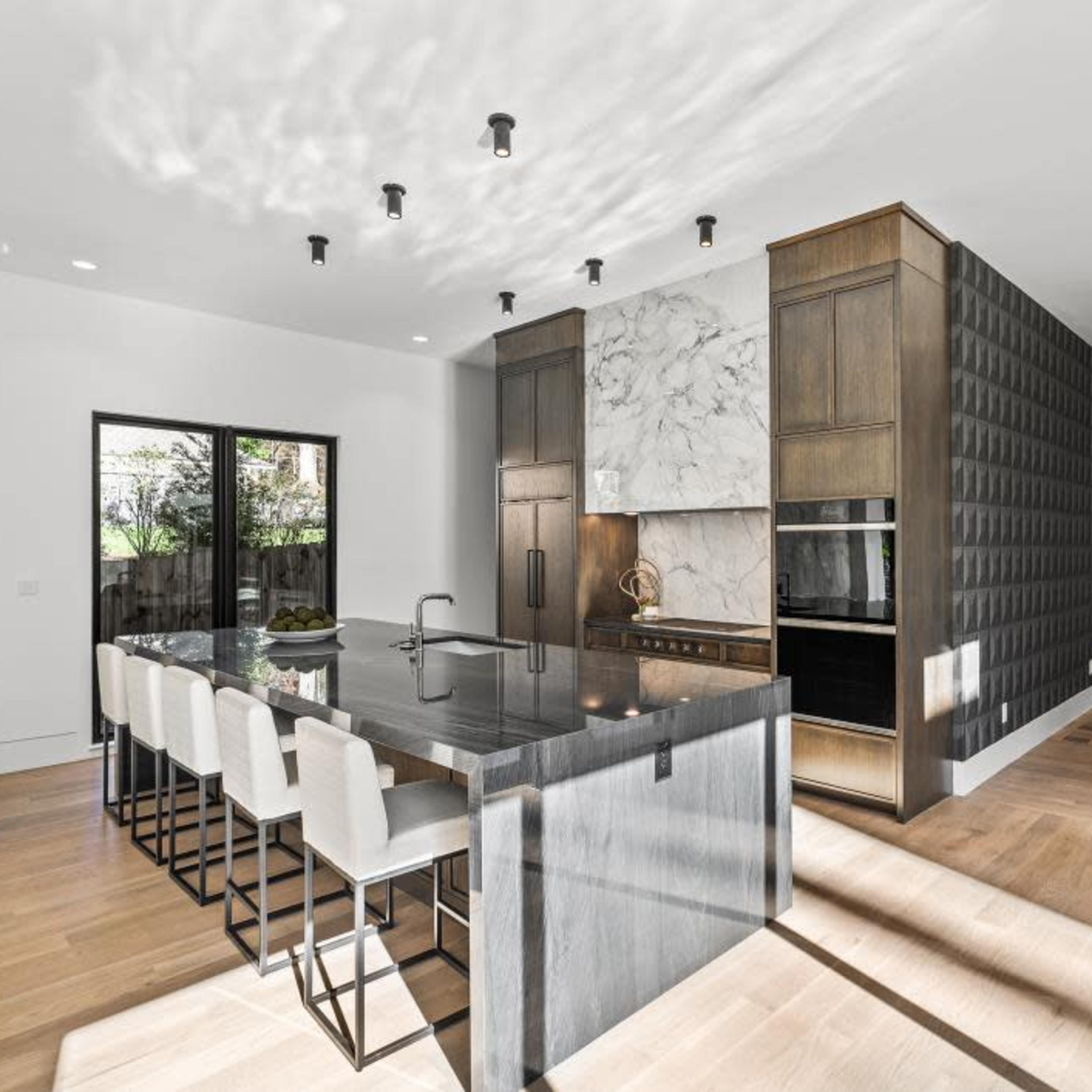 A modern kitchen with a large island, high-backed stools, and elegant cabinetry, adjacent to a living area with minimalist decor.
