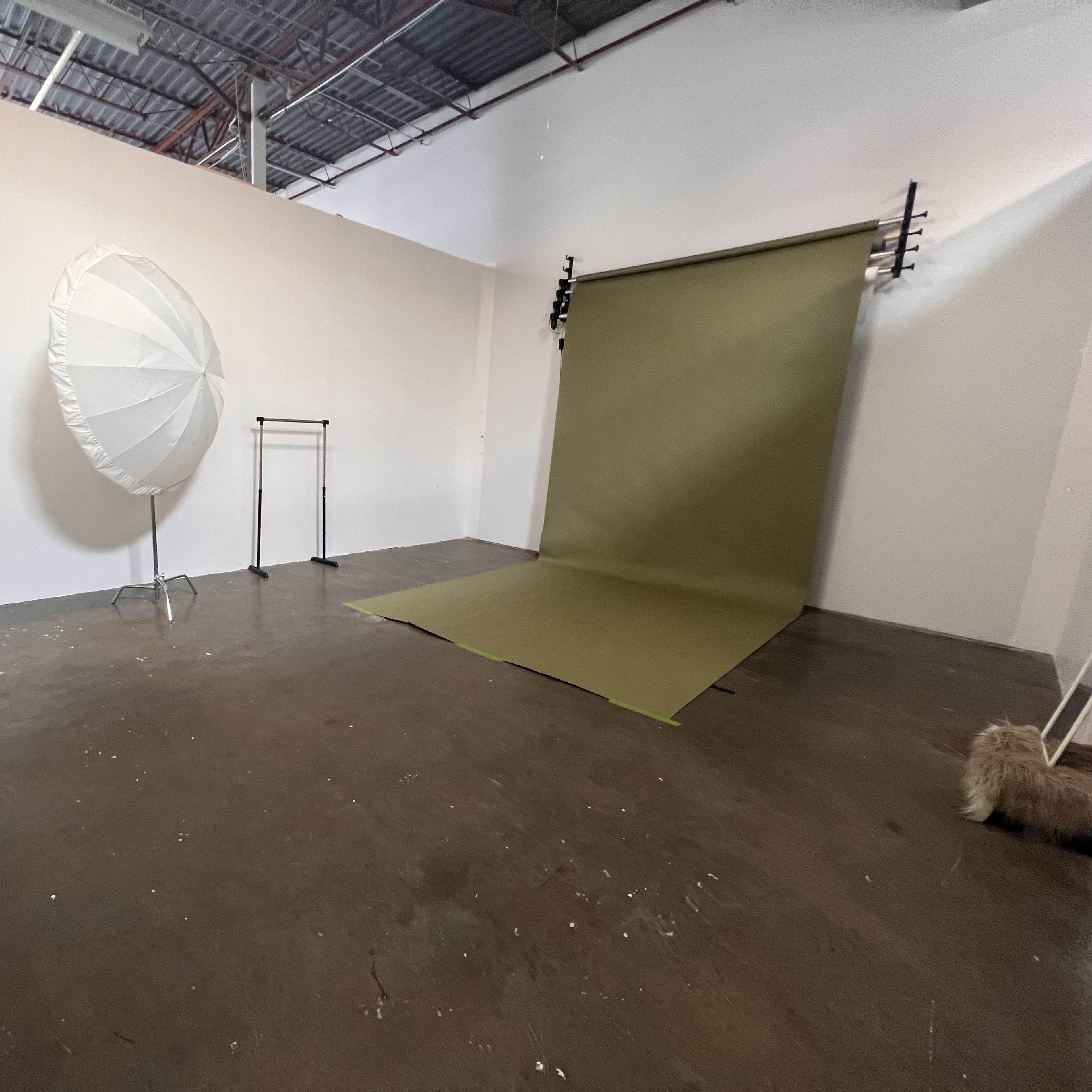A minimalist photography studio featuring a green backdrop, a large white reflective umbrella, a clothing rack, and a small table with a chair.