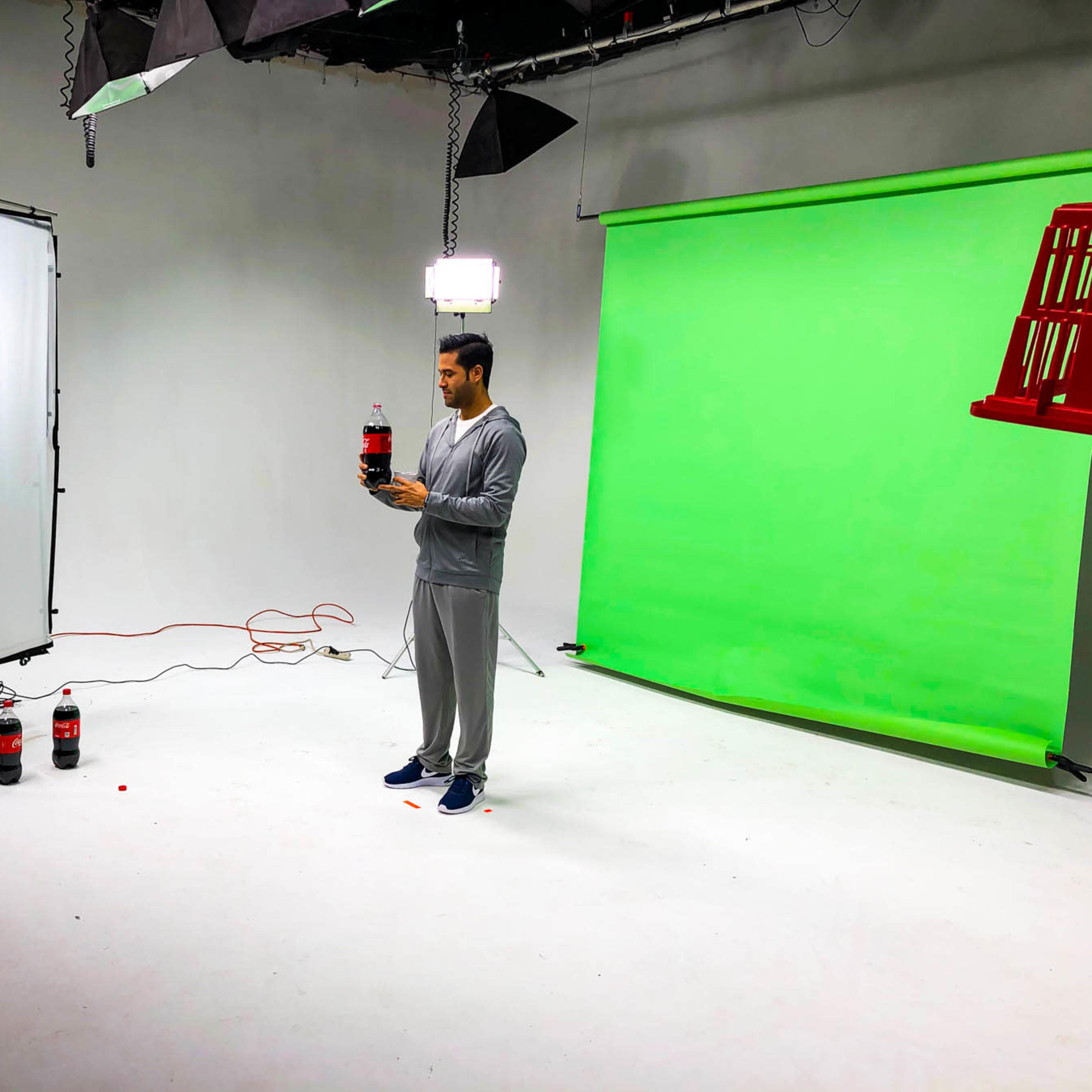 A person stands in a studio with a green screen, holding a bottle of soda, surrounded by lighting equipment and props.