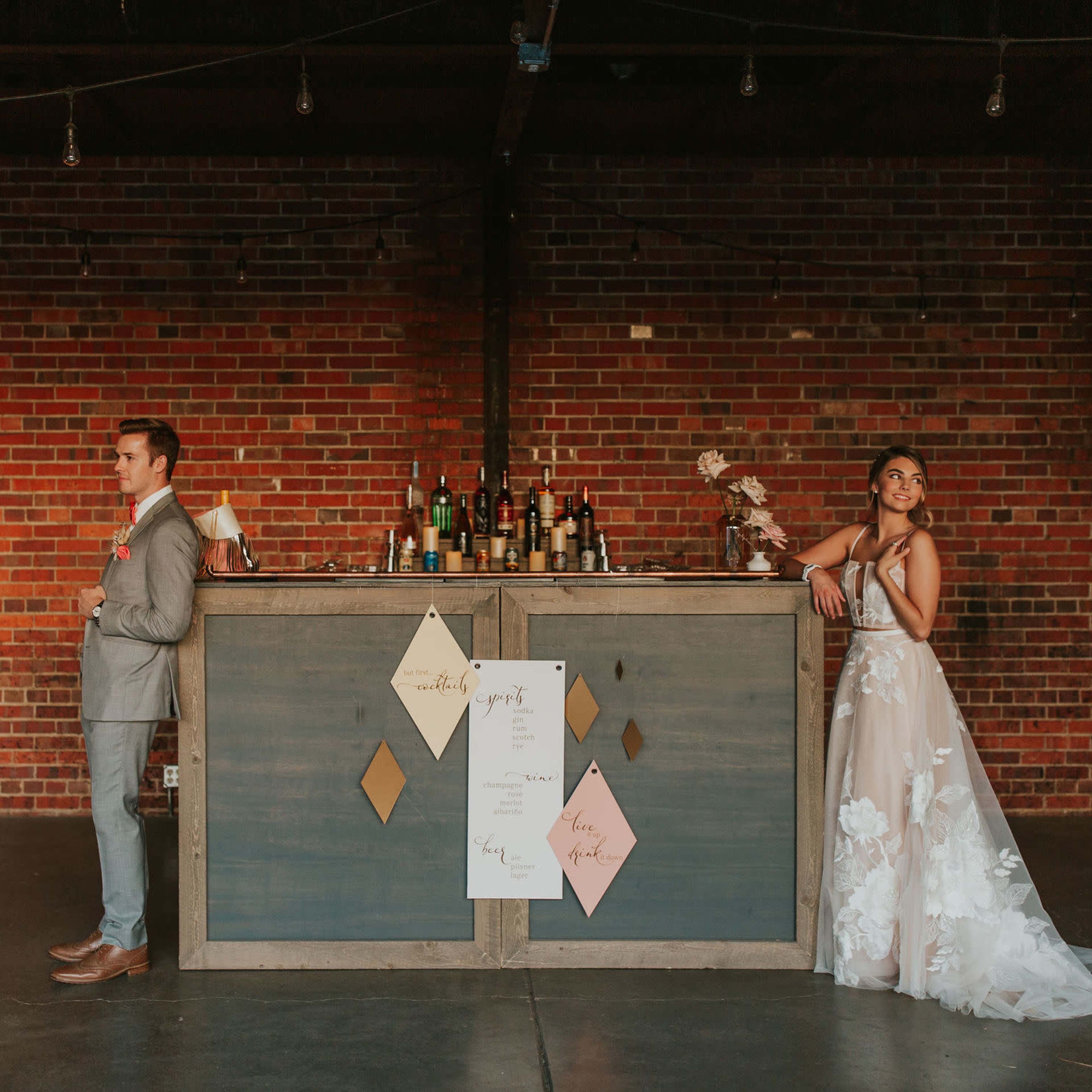 A couple stands on opposite sides of a vintage bar against a brick wall, with wedding decor displayed in front of them.