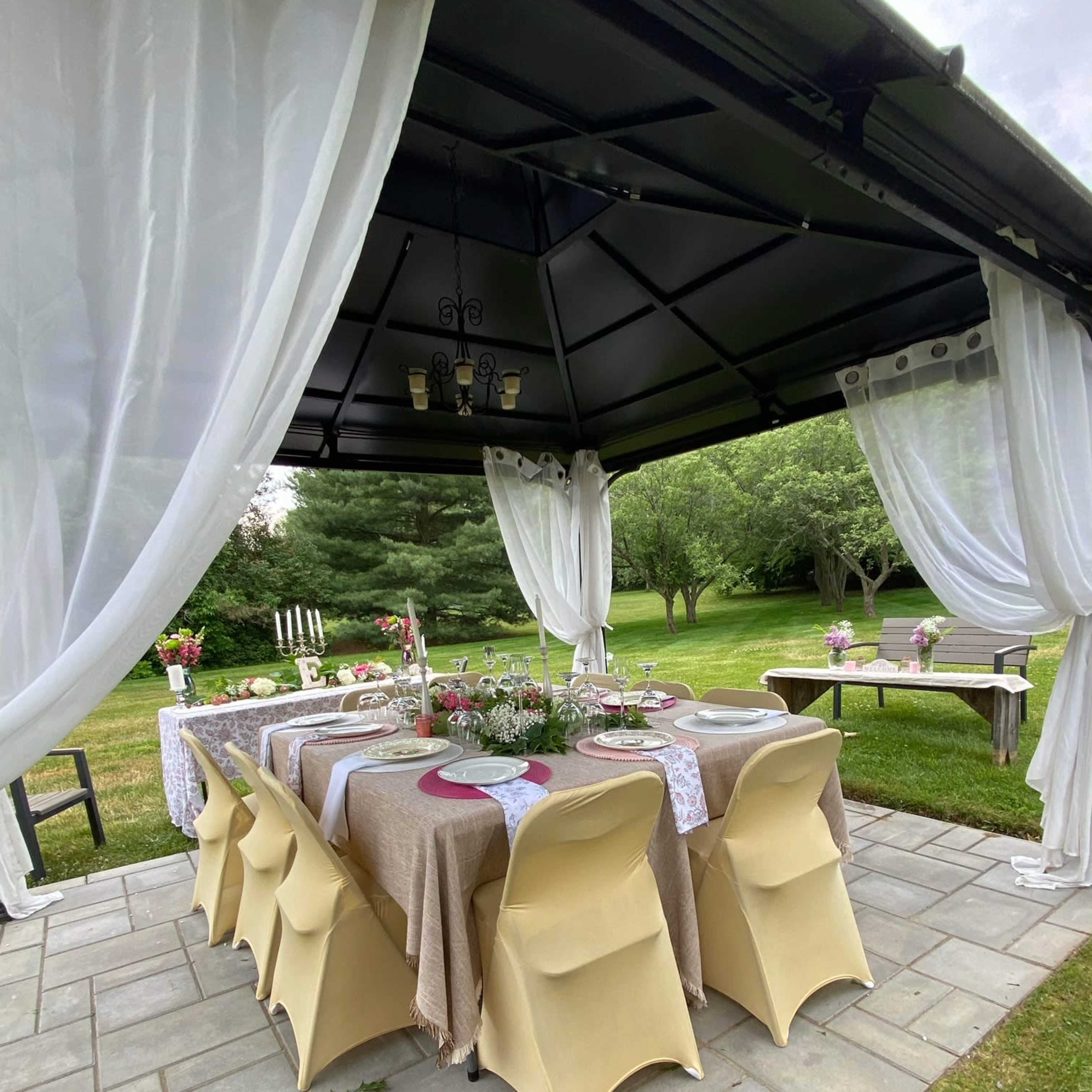 A covered outdoor dining area features a long table set with tableware and decorations, surrounded by grass and trees.