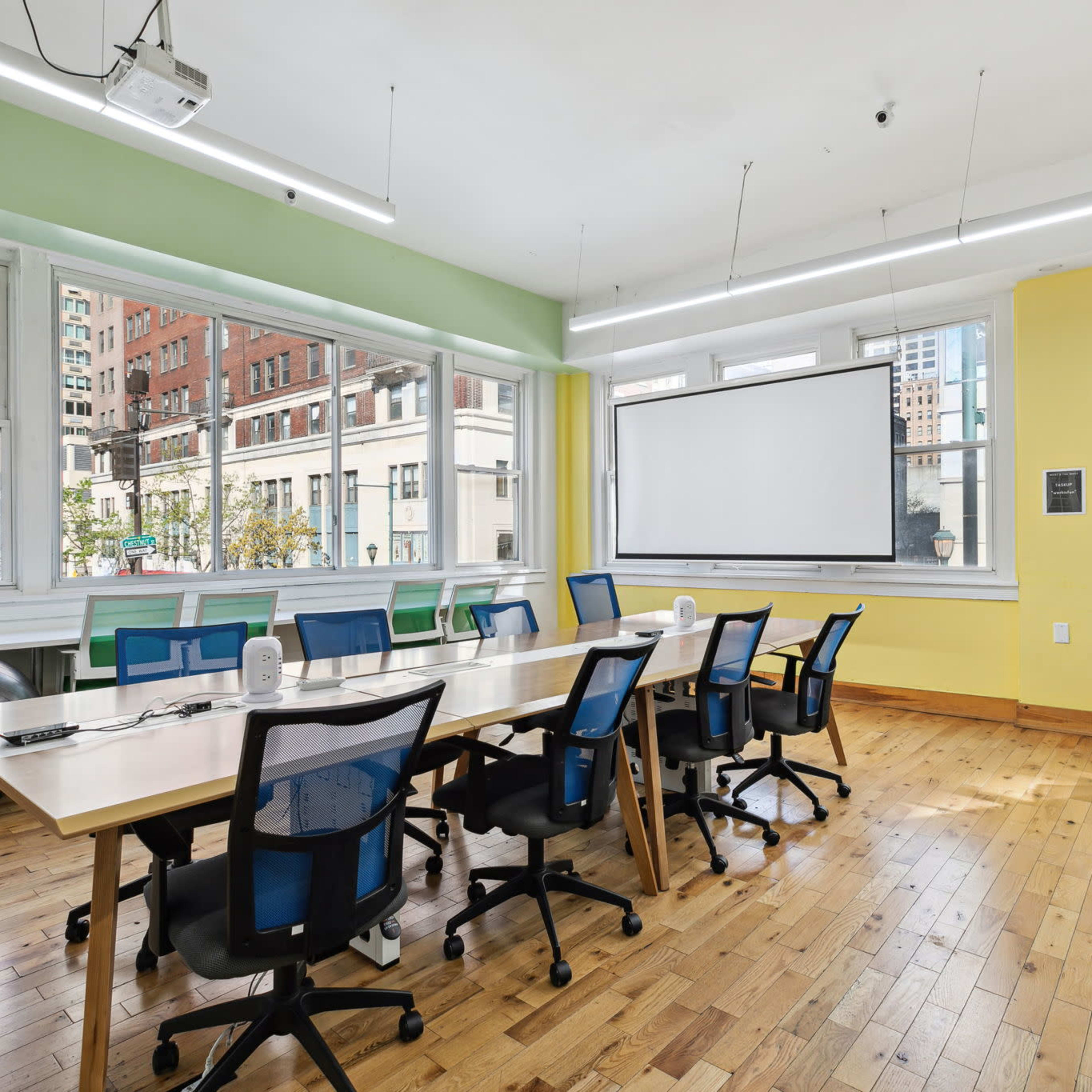 A bright, modern office space features a long wooden table surrounded by ergonomic chairs, with large windows providing city views and a projector screen at one end.