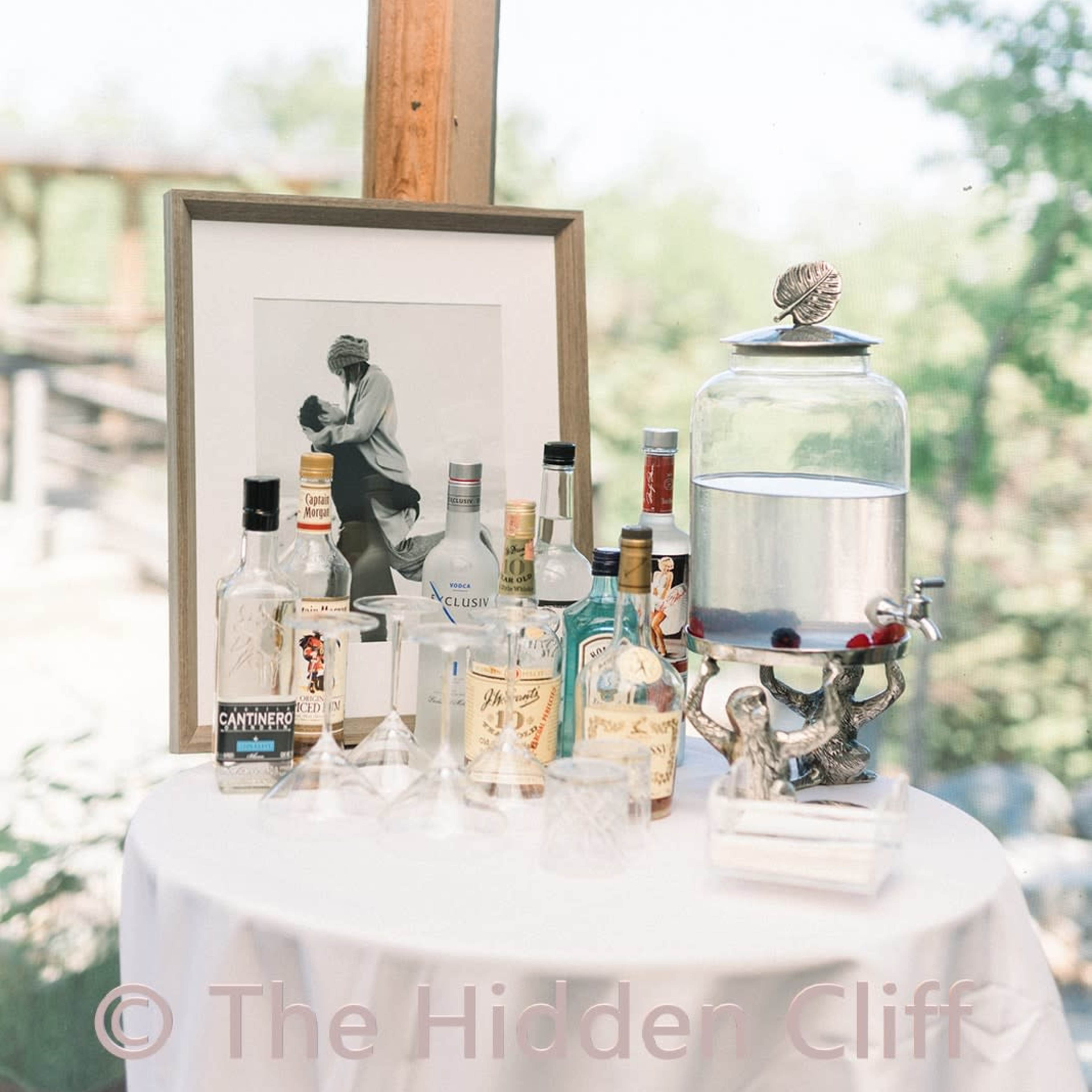 A table draped with a white cloth displays various bottles of liquor alongside a framed black-and-white photograph and a glass beverage dispenser.