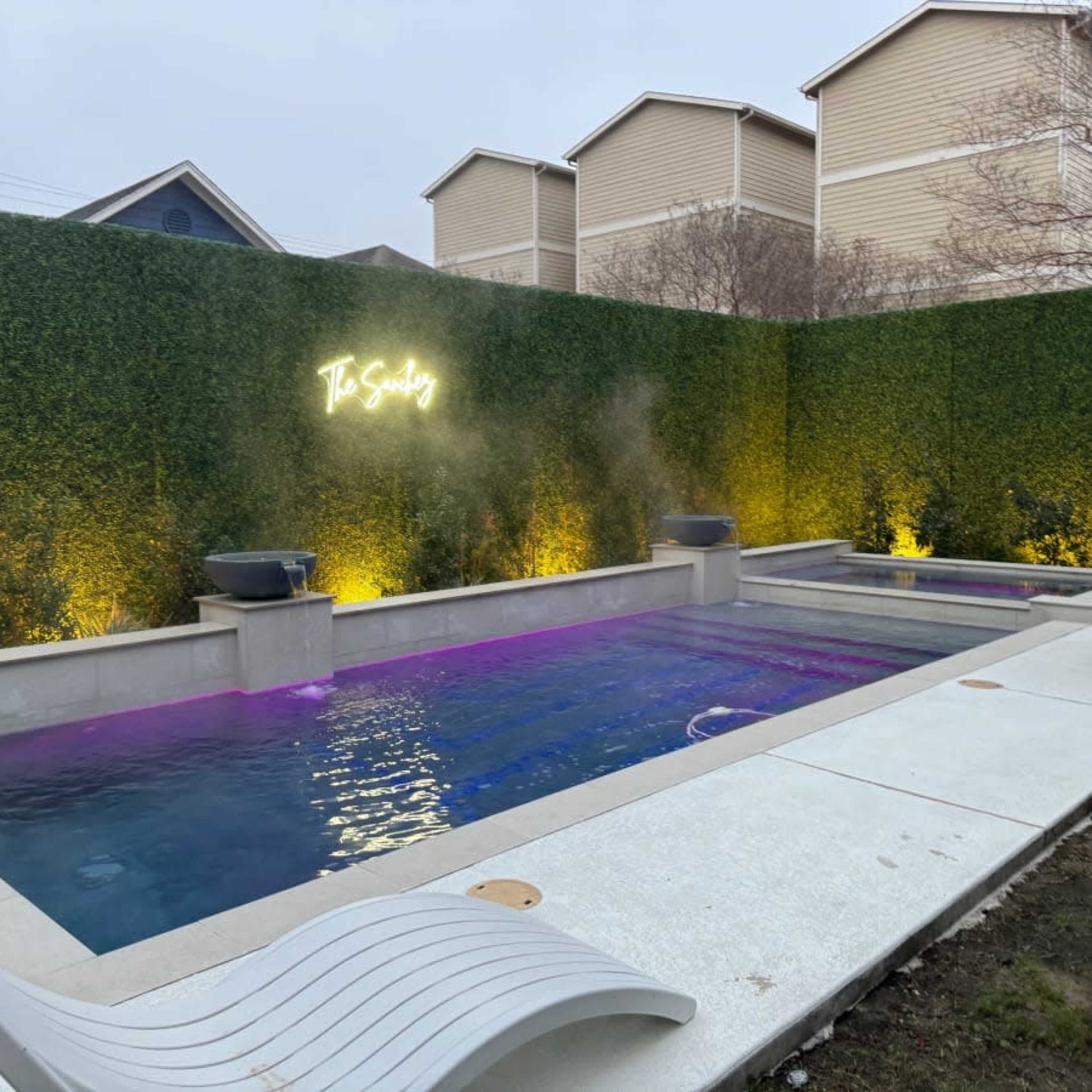 A modern outdoor pool area with two pools surrounded by a tall green hedge and illuminated by yellow lights.