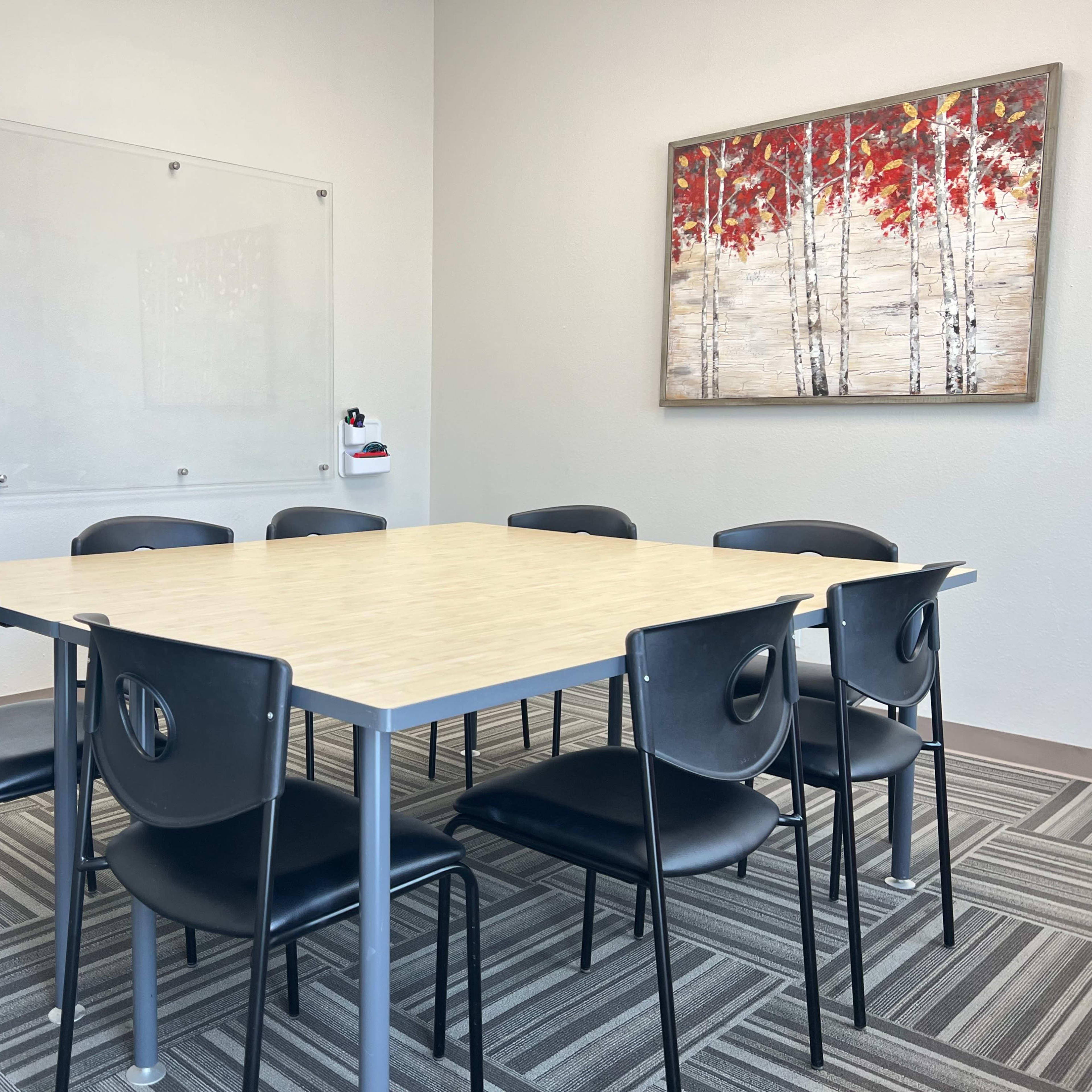 A conference room features a square table surrounded by eight black chairs, with a whiteboard and a framed artwork of trees with red leaves on the wall.