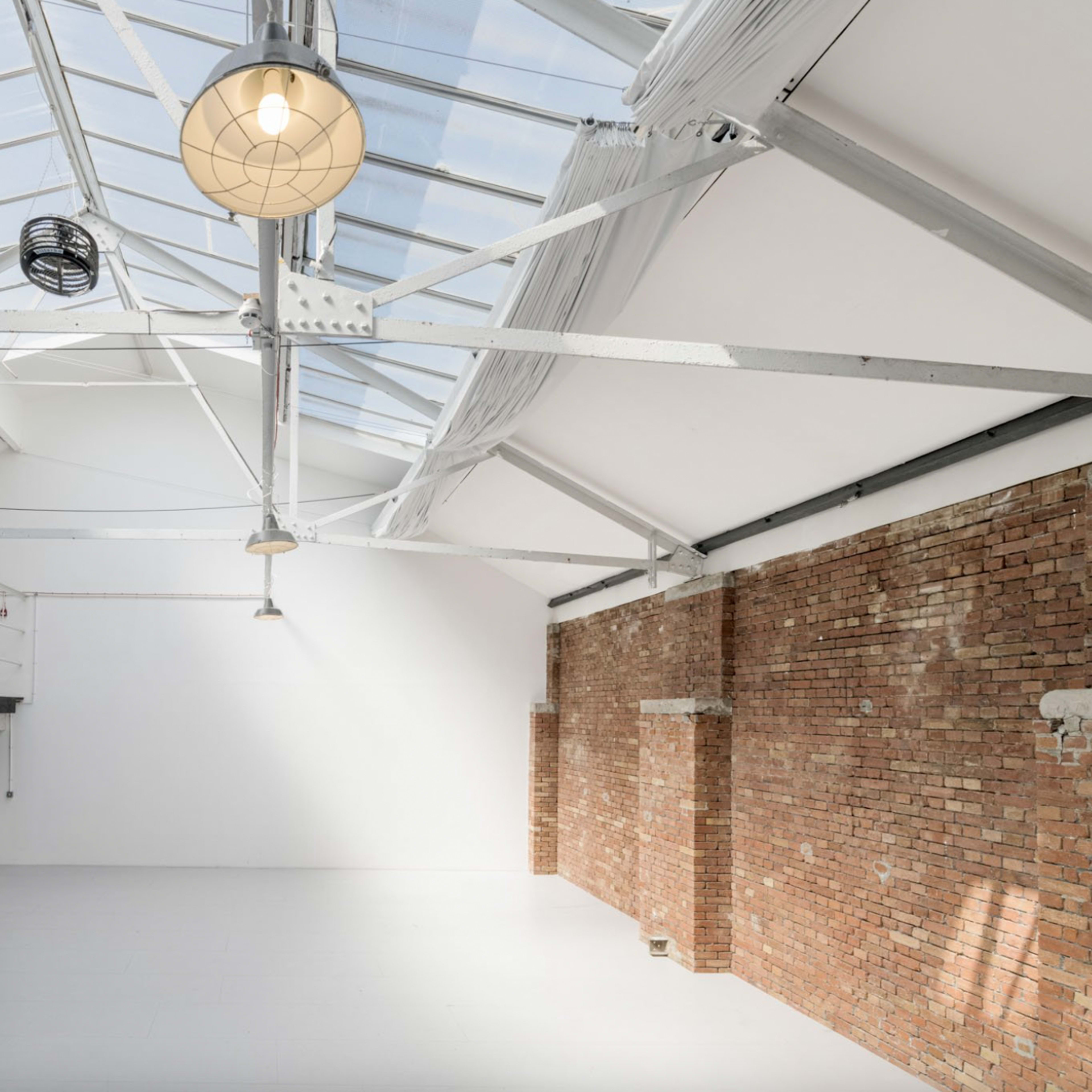 A spacious room with a high ceiling, featuring large windows, exposed brick walls, and minimal furnishings.