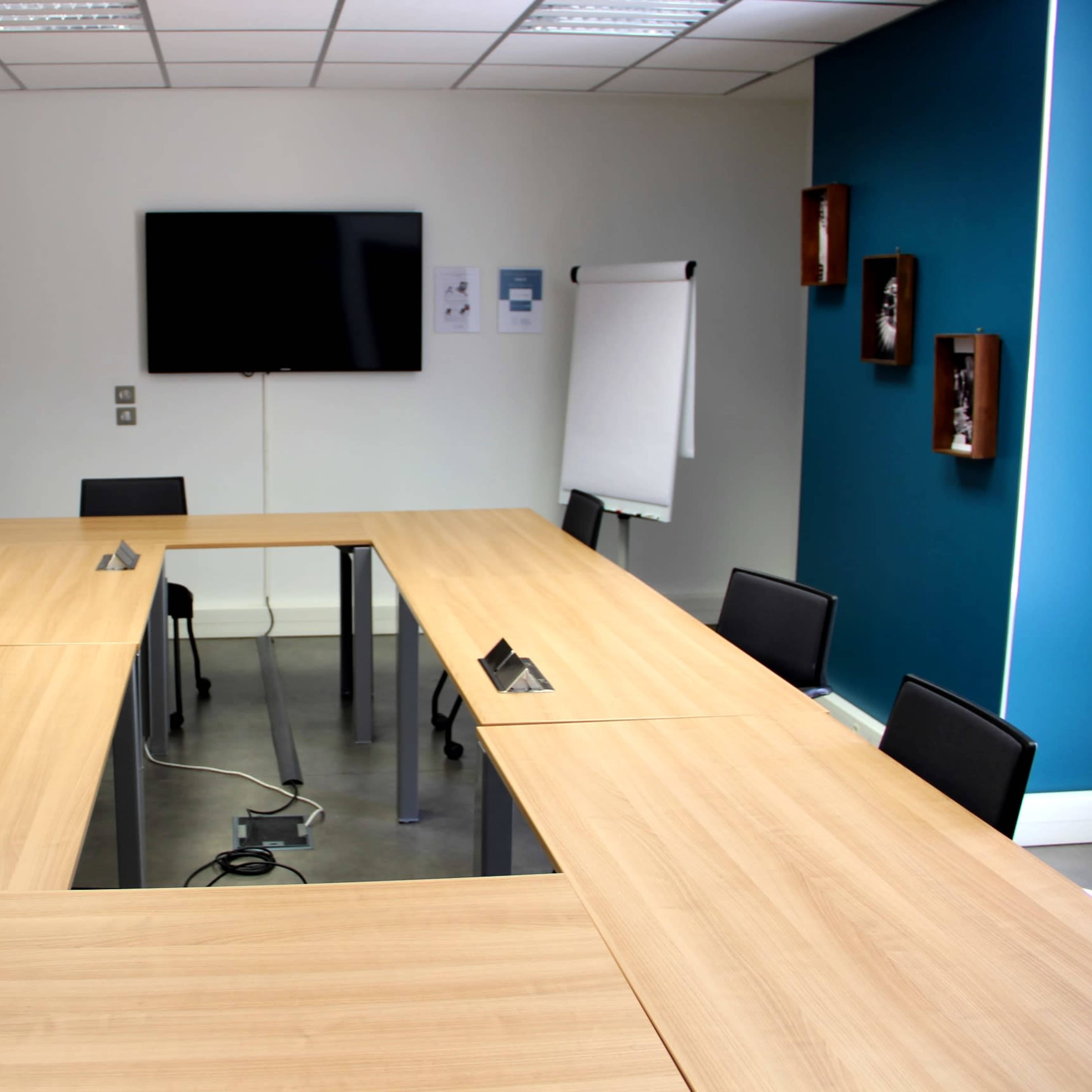 A modern conference room features a long wooden table, several black chairs, a wall-mounted television, and a whiteboard in a bright professional setting.