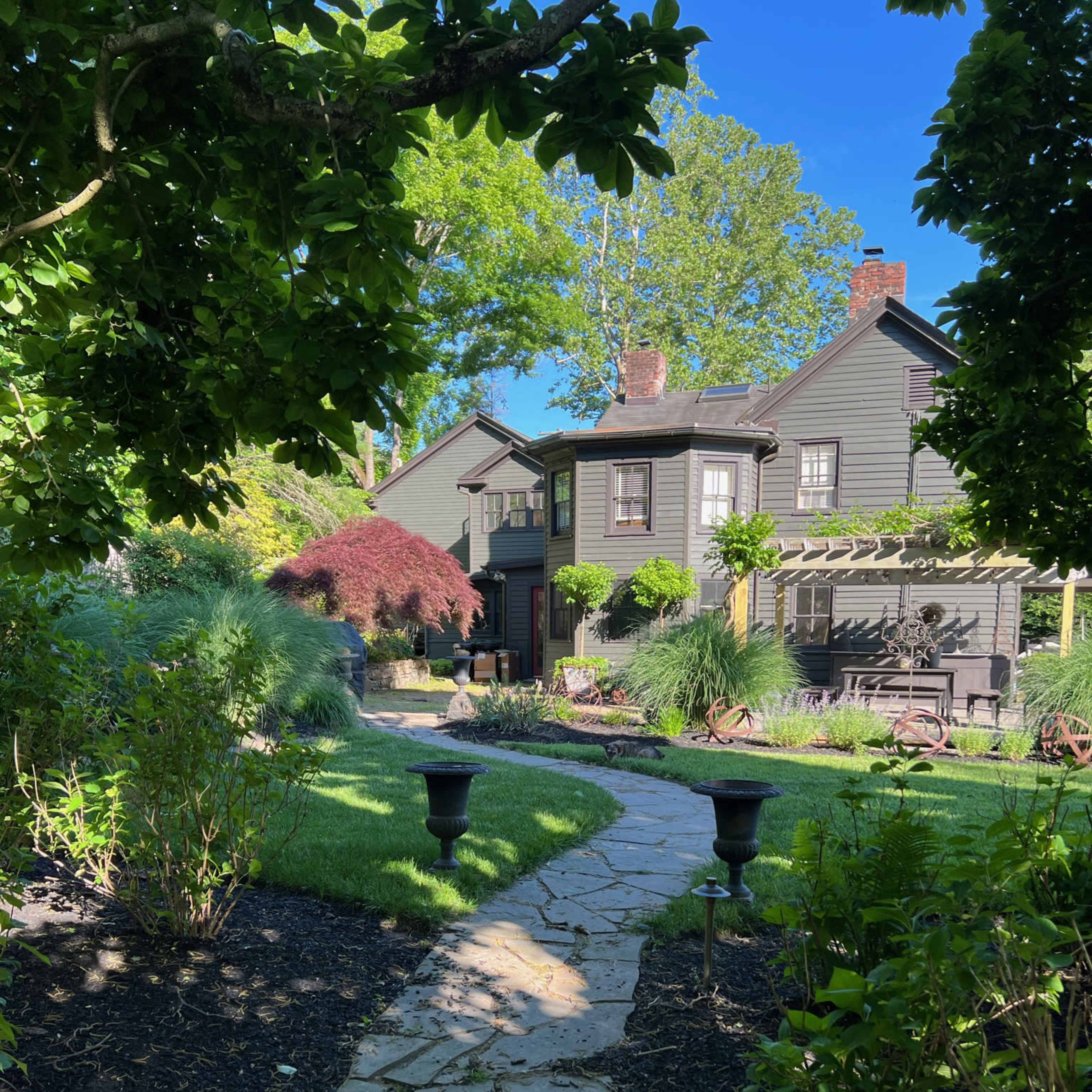 A stone path leads through a landscaped garden to a dark-colored house with multiple stories and a porch.