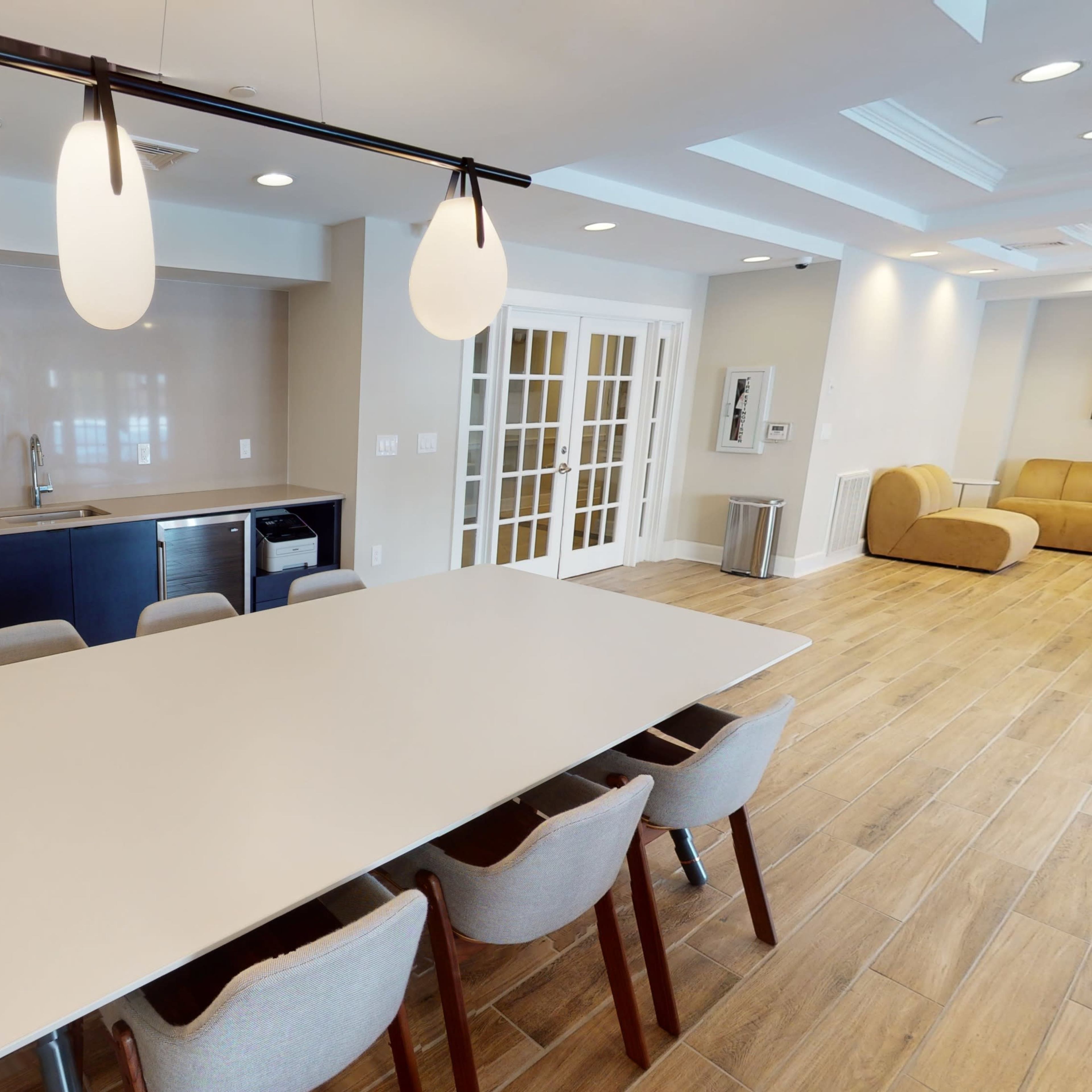 A modern dining area with a long table surrounded by upholstered chairs, a kitchenette with appliances, and decorative elements against a neutral-colored wall.