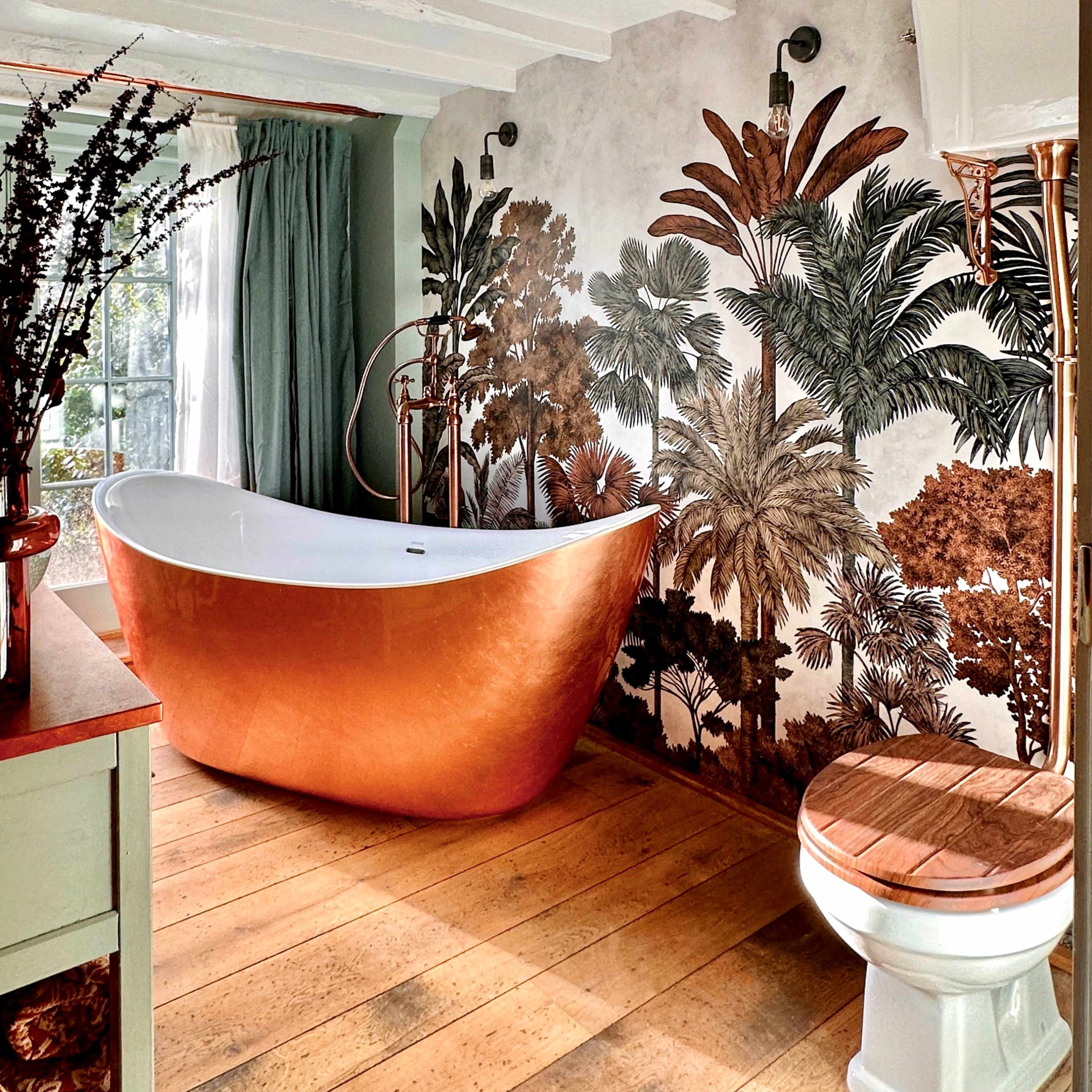 A stylish bathroom with a copper freestanding bathtub, a decorative mural of tropical plants on the wall, and wooden flooring.