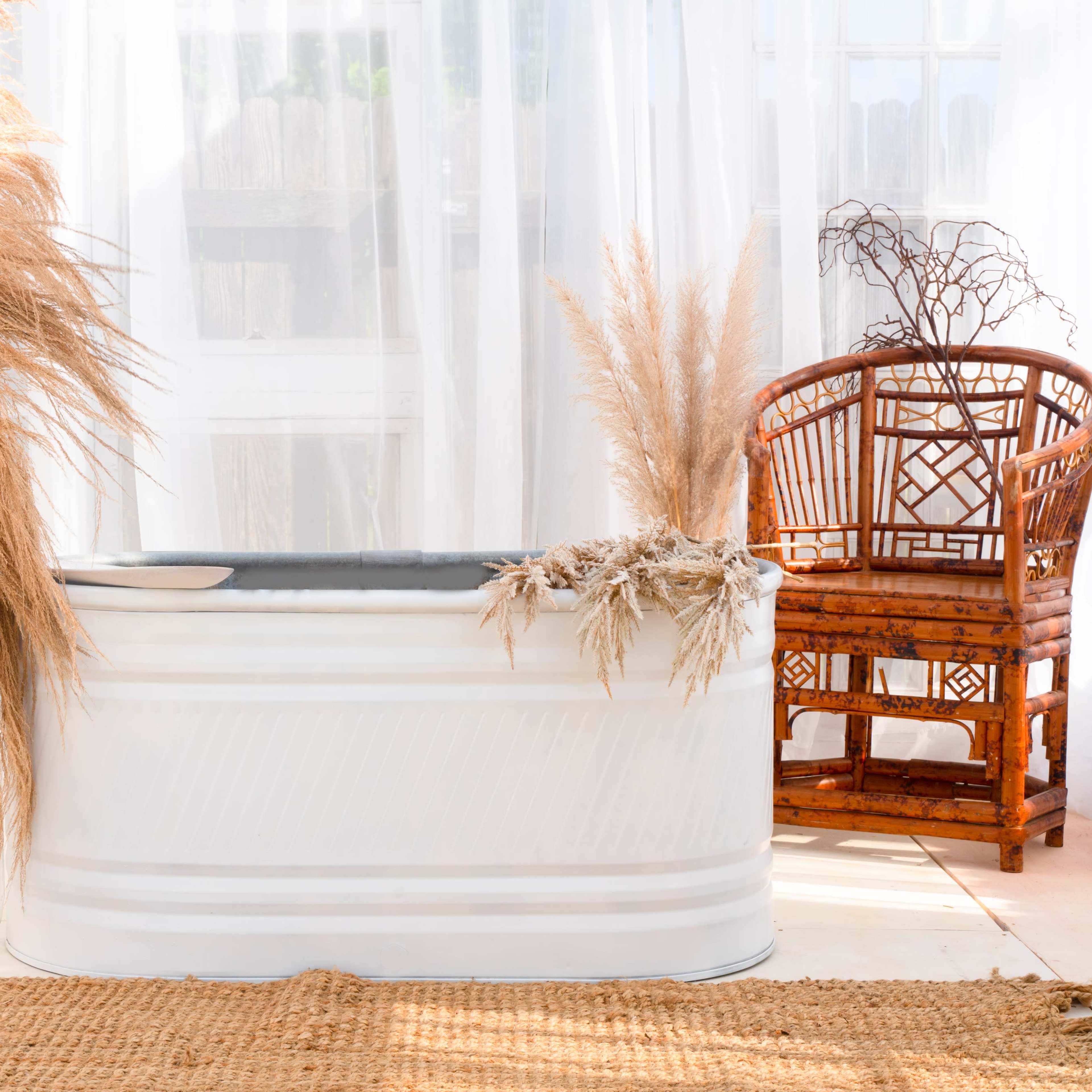 A white bathtub accompanied by dried pampas grass and a wicker chair, set against a backdrop of sheer curtains and a light-colored floor.