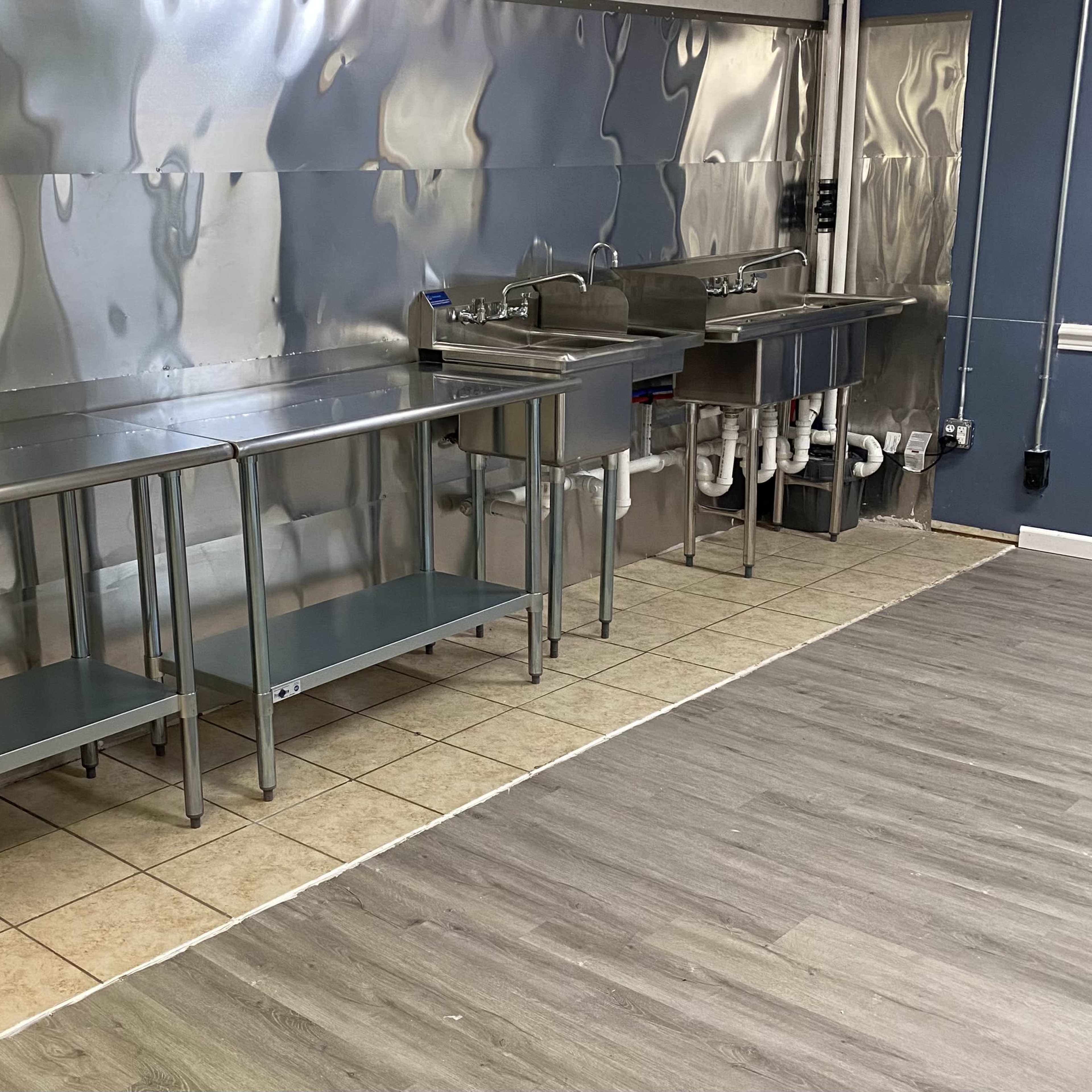 A commercial kitchen space featuring stainless steel work tables and sinks against a metal wall with tiled flooring.