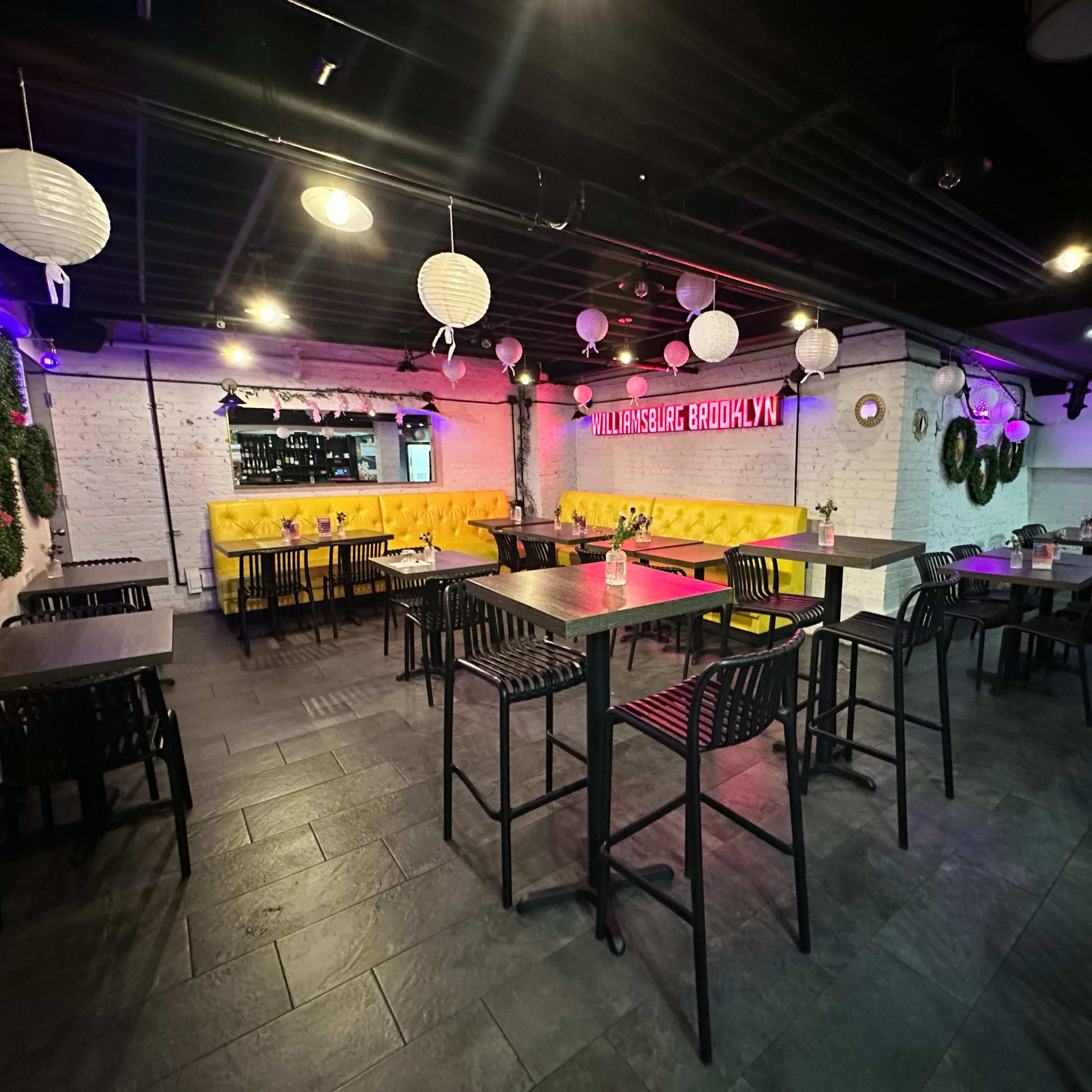 A dimly lit restaurant interior with black tables and chairs, colorful decor, and a bright neon sign on the wall.
