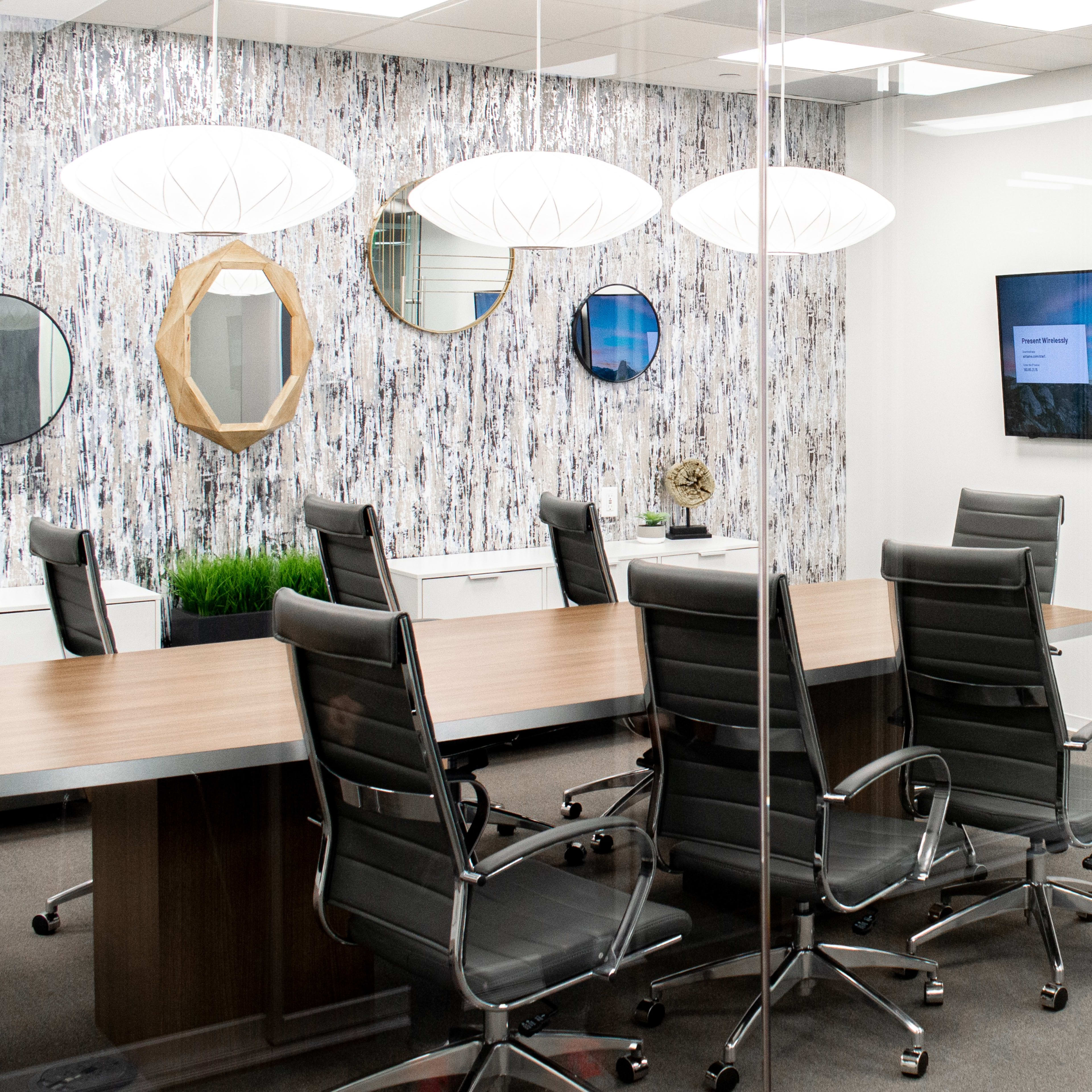 A modern conference room with a long wooden table, black leather chairs, mirrored wall decor, and two screens displaying presentations.