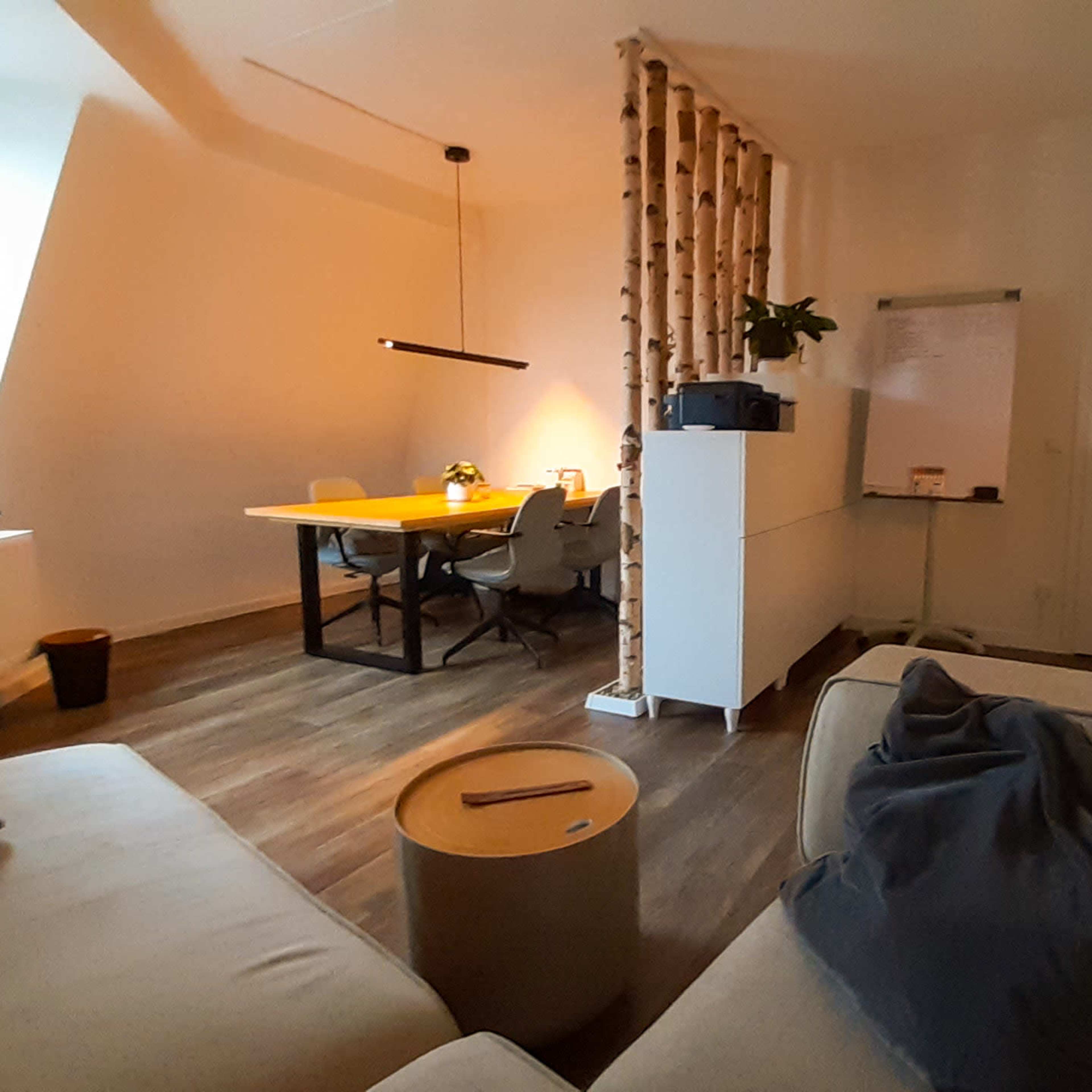 A modern room features a light-colored sofa, a wooden dining table with chairs, and a partition made of wooden beams, along with a whiteboard and potted plants.