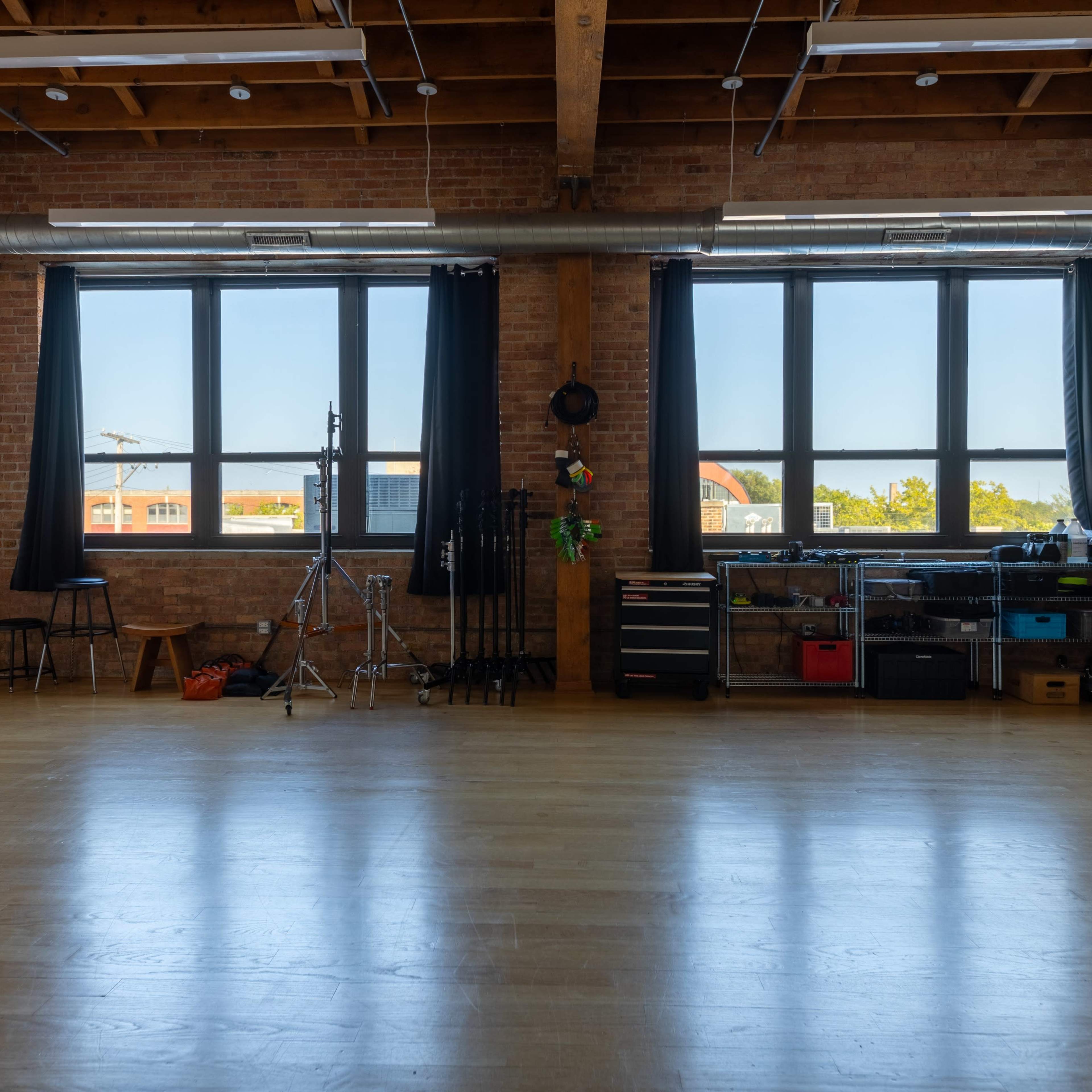 An empty studio space with large windows, wooden beams, and various photography equipment organized along one wall.