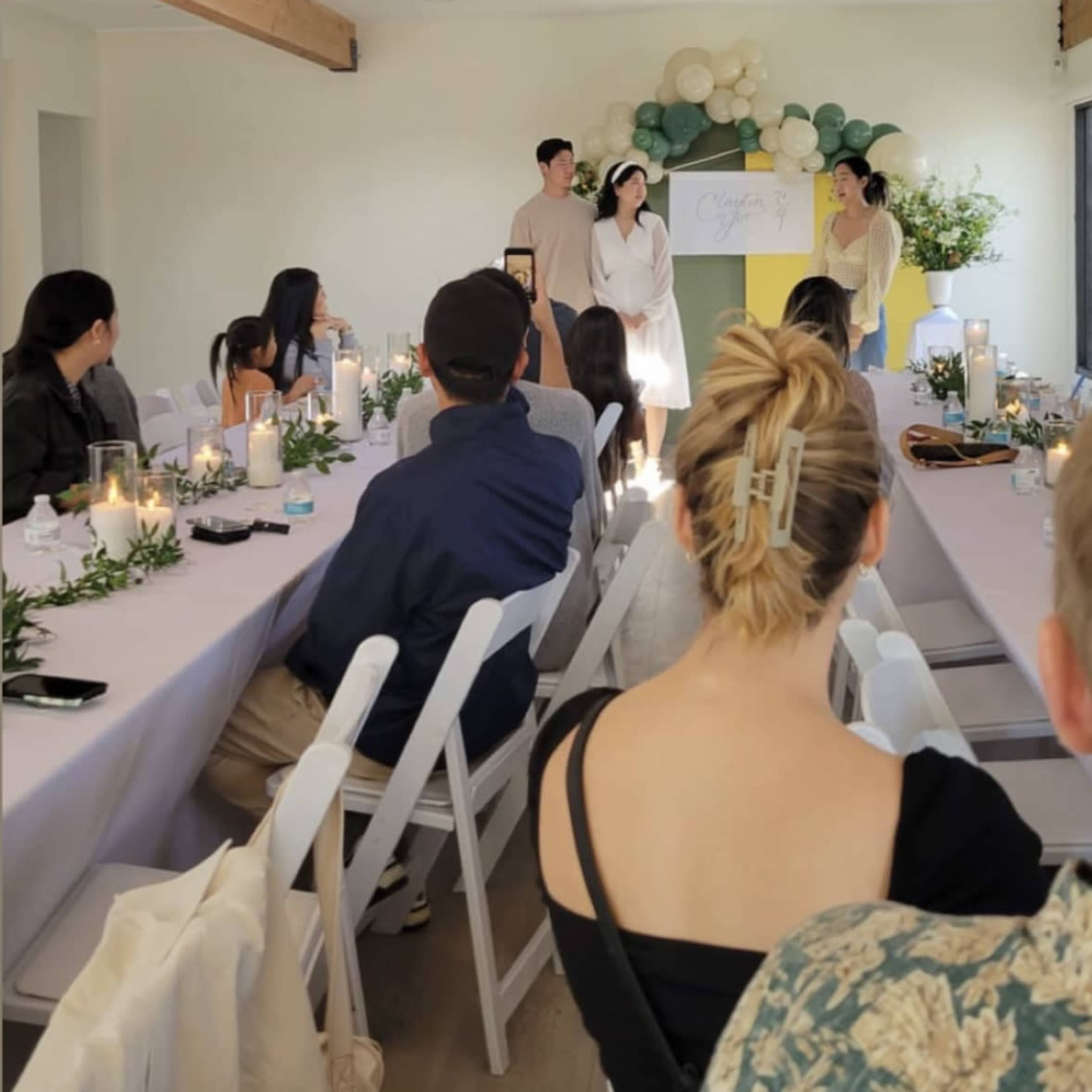 A group of people is seated at a long table in a well-lit room with wooden beams, while two individuals stand at the front, presenting something.