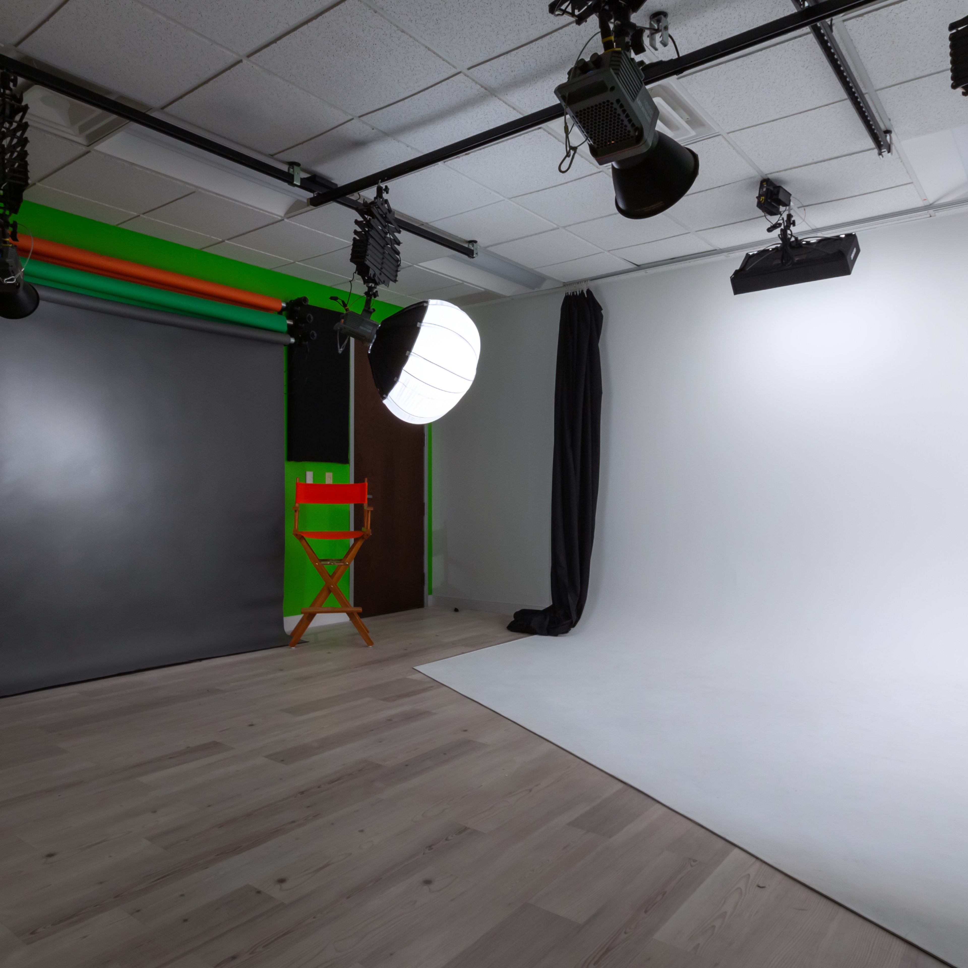 A photography studio with a gray backdrop, a lit area with a white backdrop, two director's chairs, and various lighting equipment.