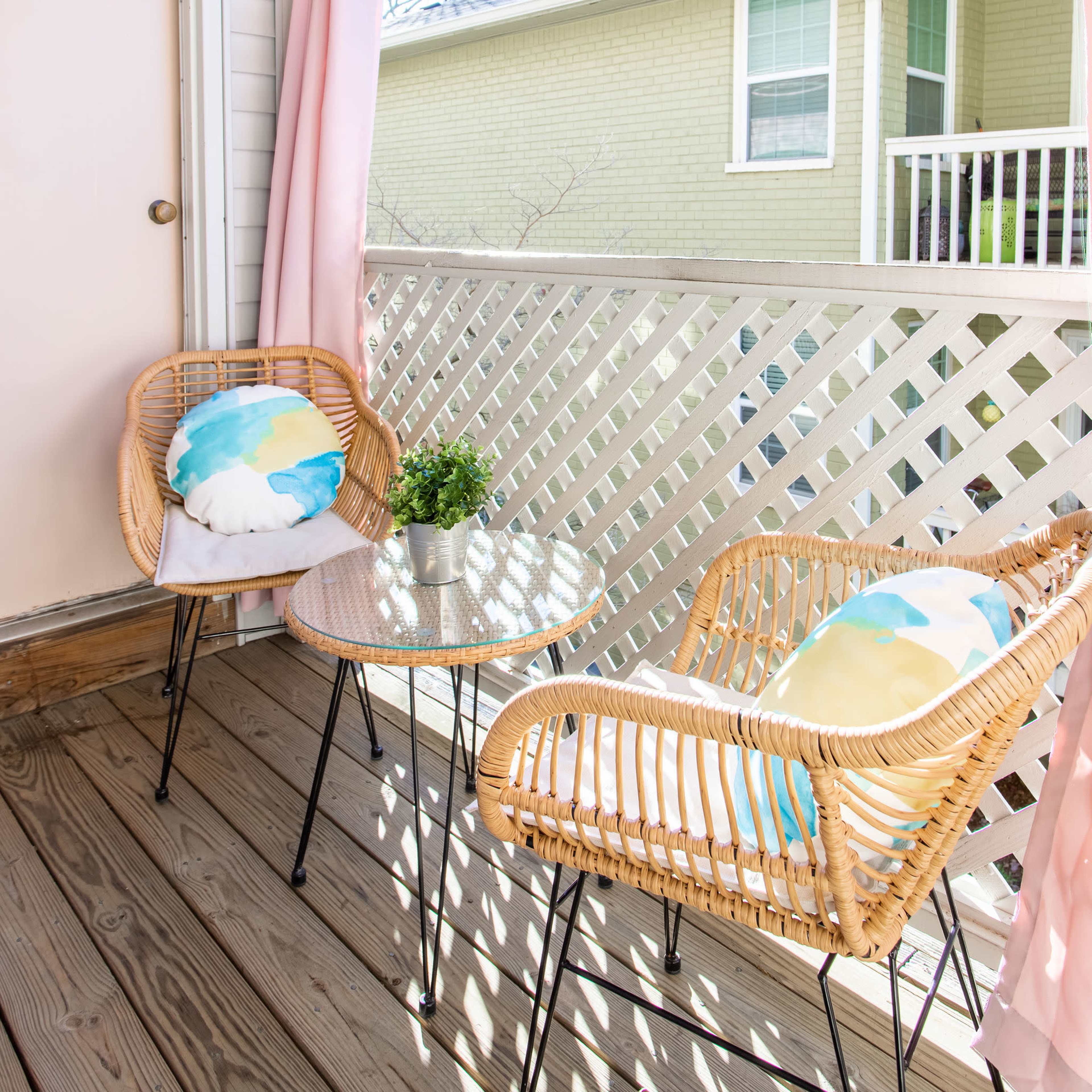A small balcony features two wicker chairs with colorful cushions and a glass table, surrounded by a wooden railing and light-colored walls.