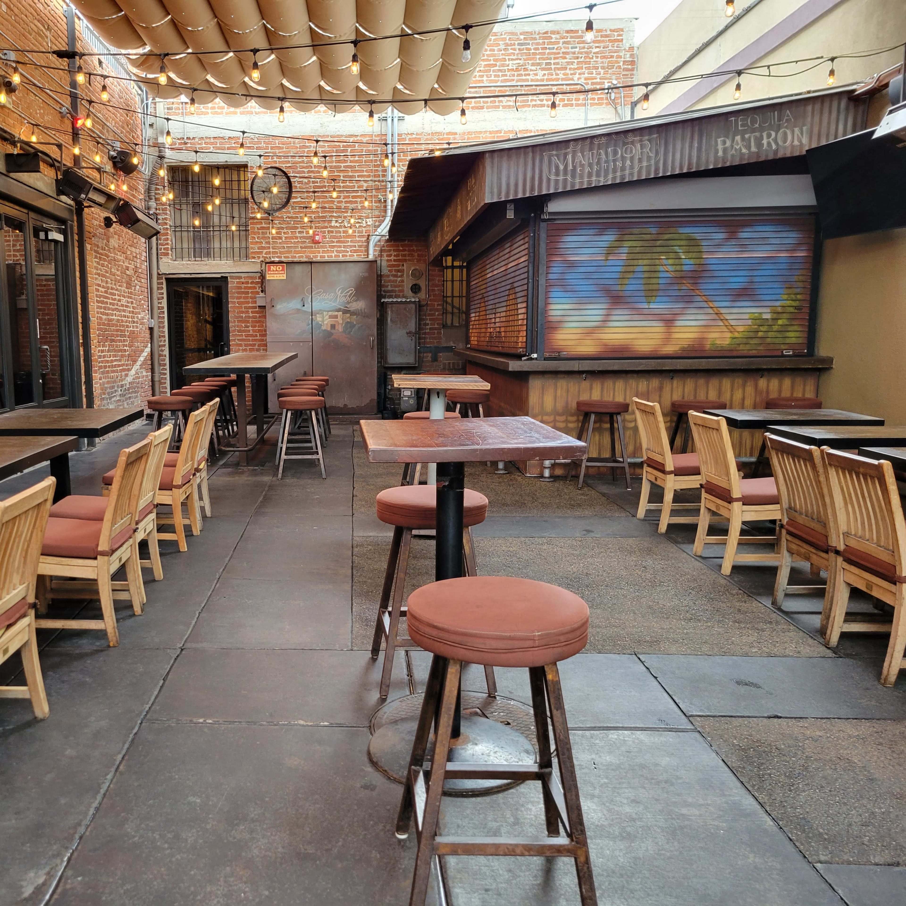 A patio area of a restaurant featuring wooden tables and chairs against a backdrop of brick walls and overhead string lights.
