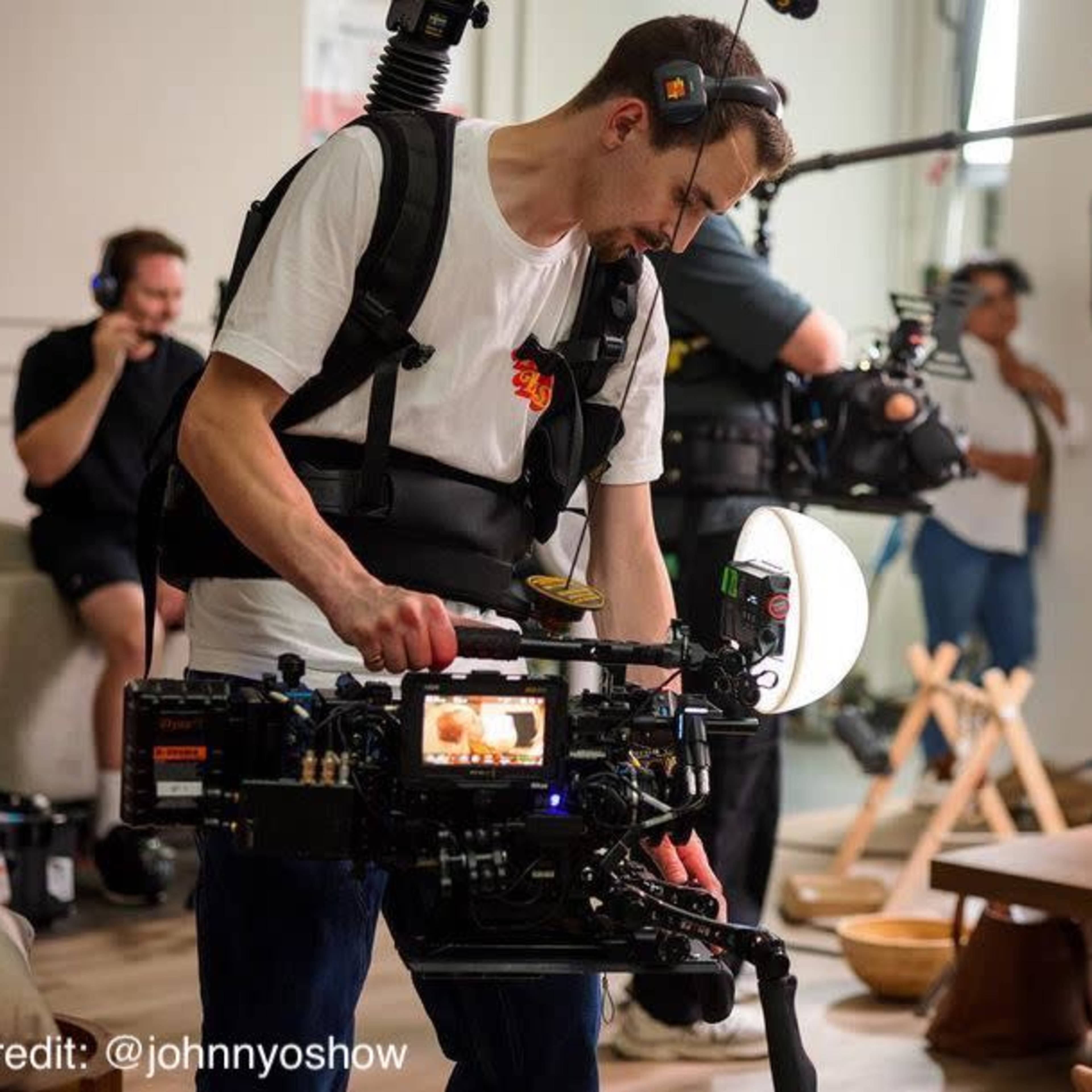 A film crew sets up in a bright room, with a cameraman using a handheld camera to film a mother holding her baby while other crew members adjust equipment.