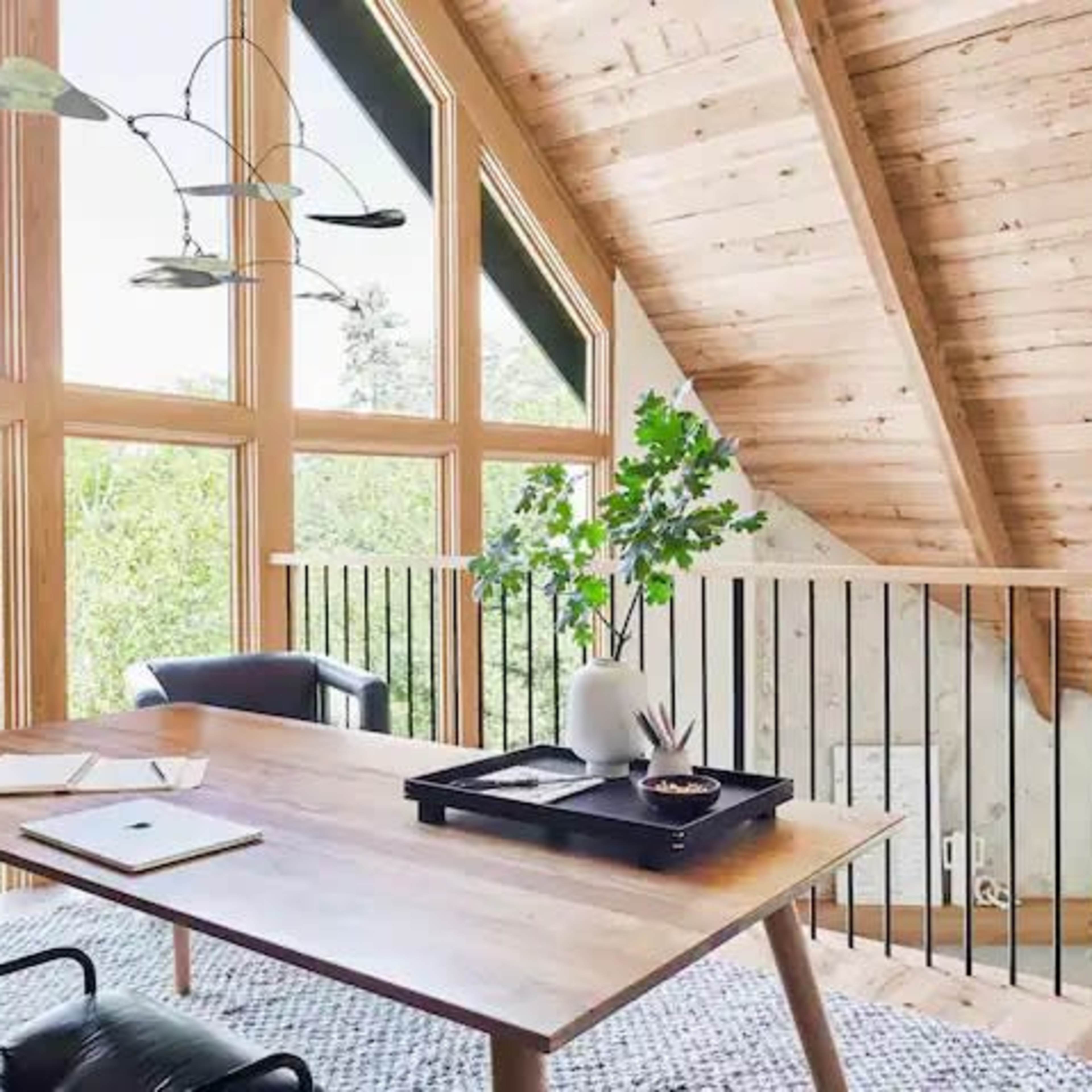 A wooden table with a notepad and pen sits in a bright room featuring large triangular windows and wooden beams overhead.