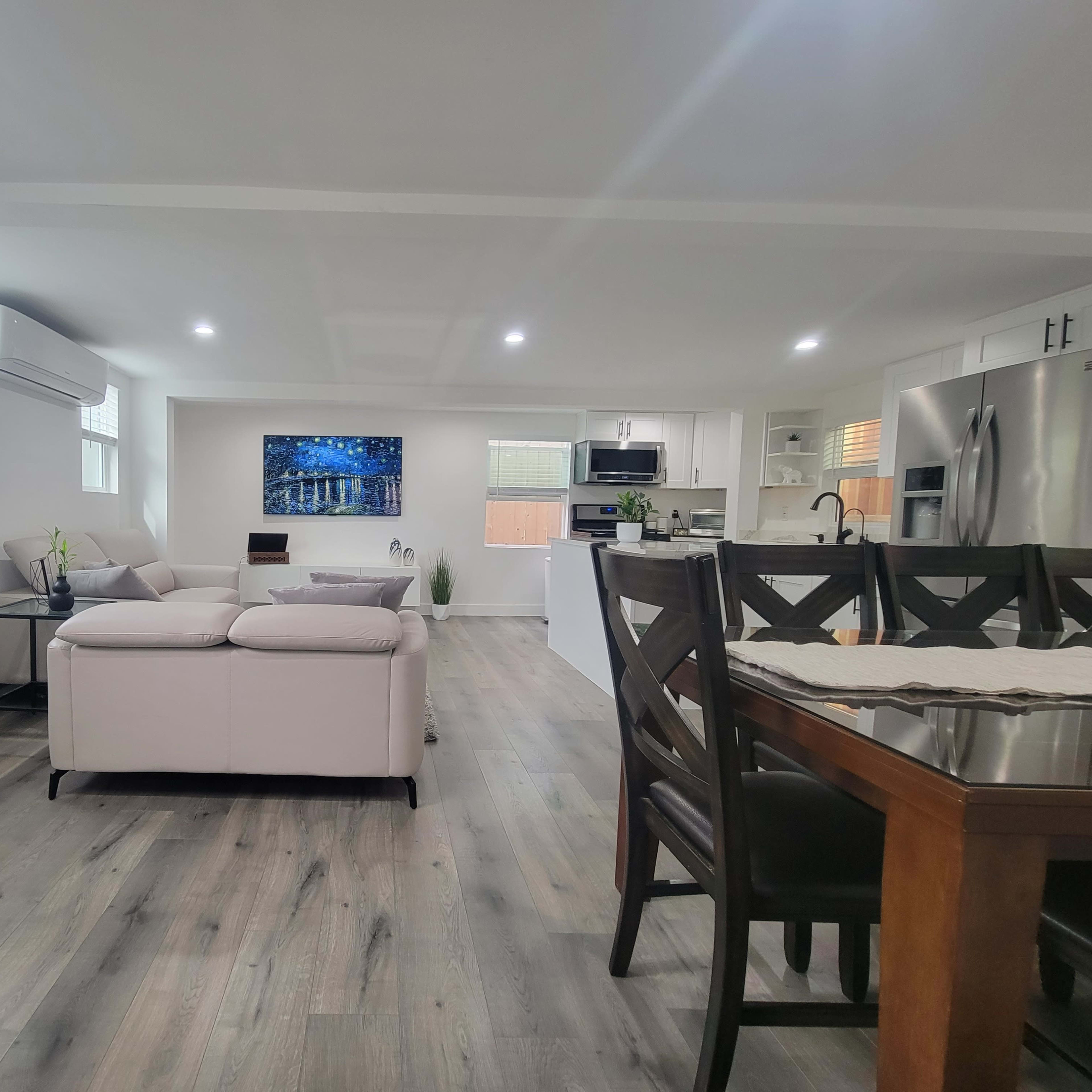 A modern, open-concept living space featuring a light-colored sofa, a dining area with a wooden table, and a kitchen in the background, all illuminated by natural light.