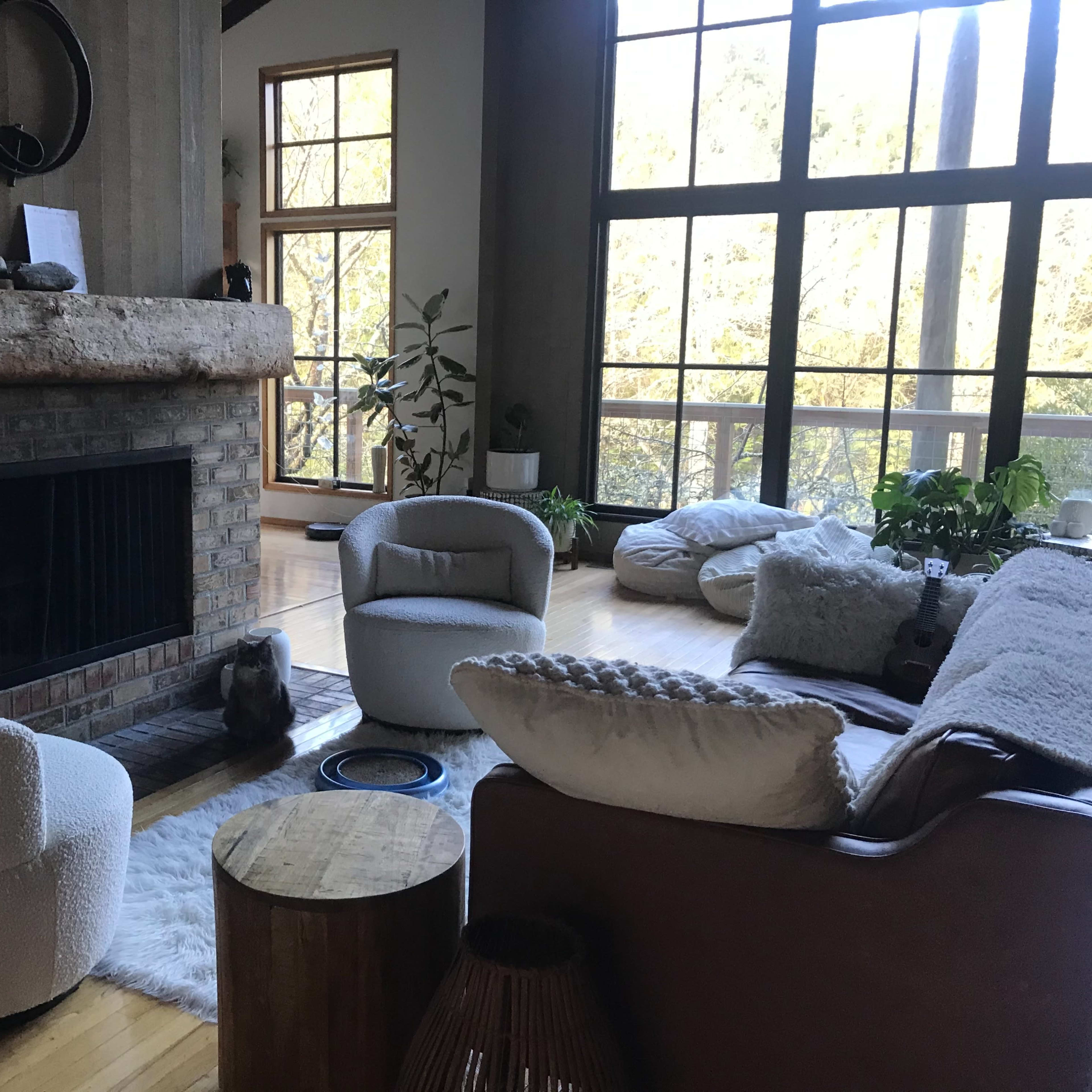 A spacious living room features a large fireplace, two round chairs, a wooden table, and large windows allowing natural light to fill the space.