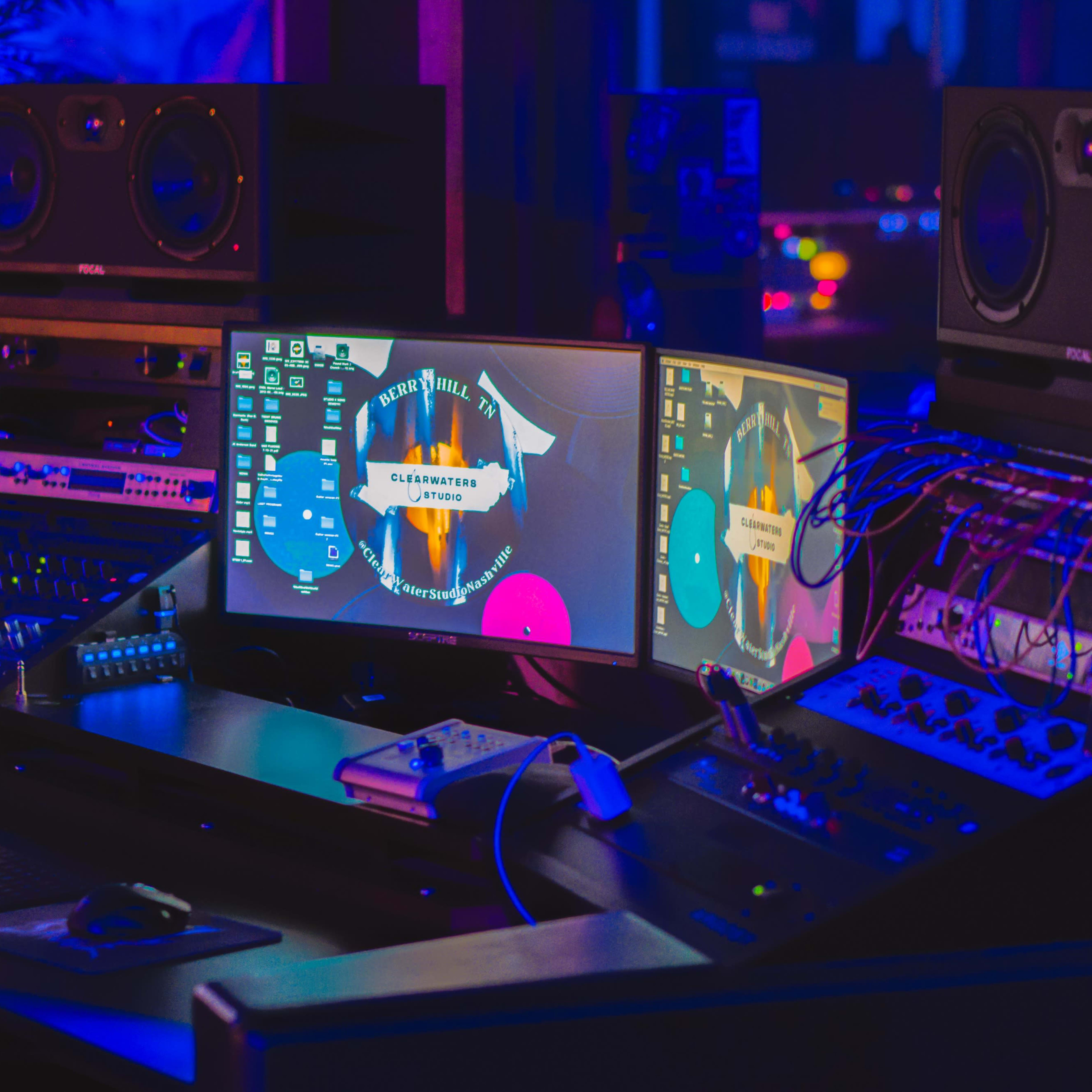 A computer workstation in a music studio features multiple monitors, audio equipment, and a digital interface amidst a blue-lit environment.