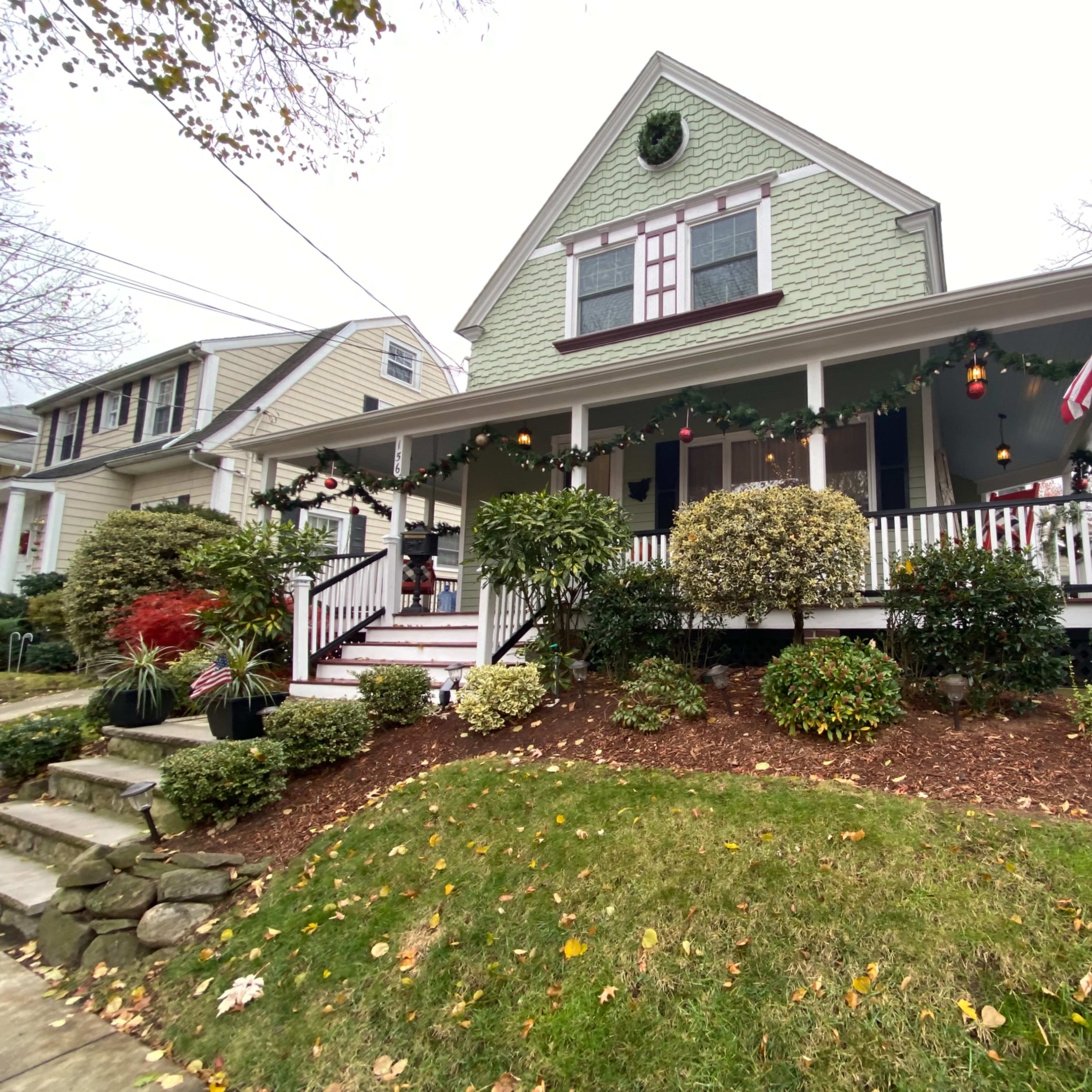 A green, two-story house with a front porch is decorated for the holidays, featuring garlands, ornaments, and an American flag.