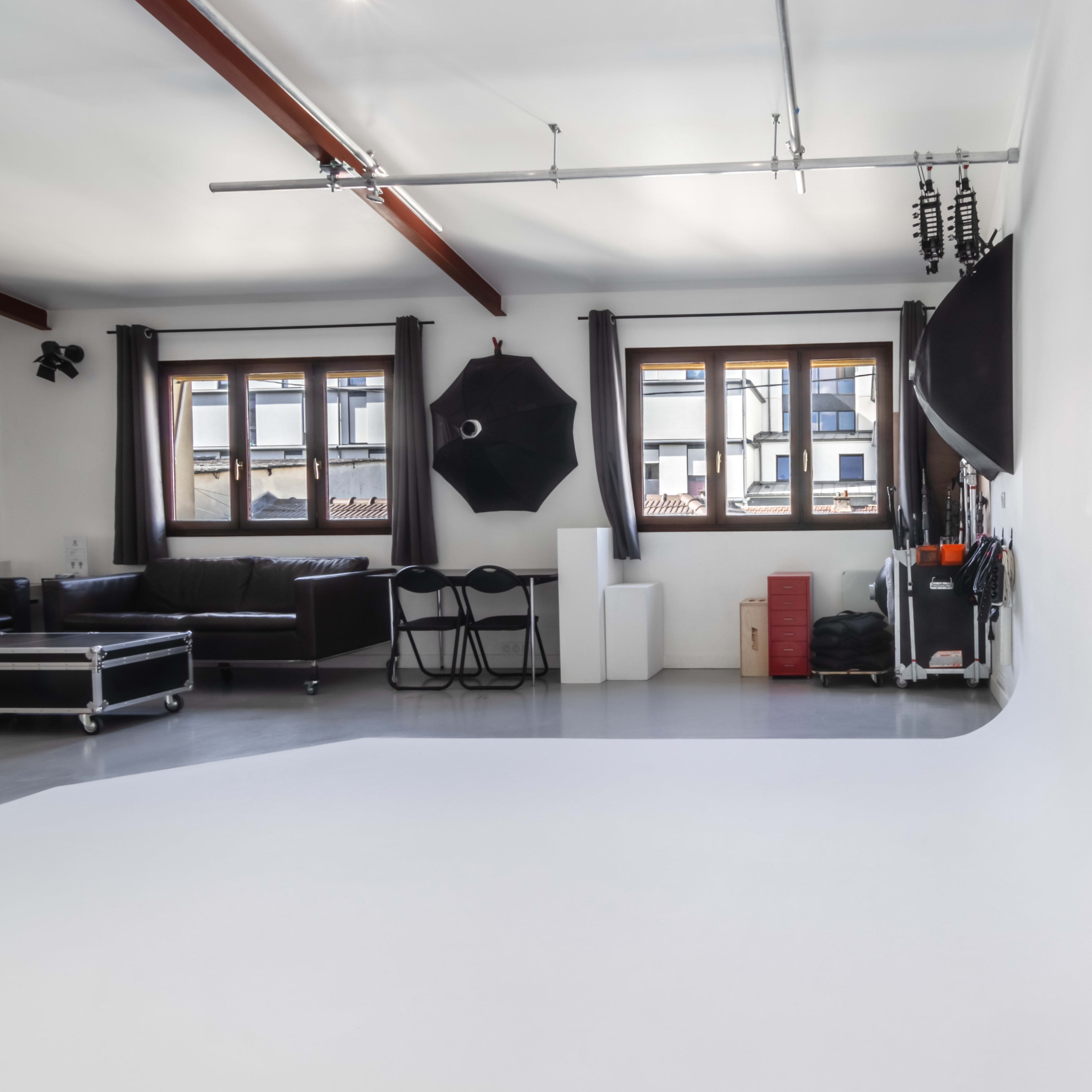 A photography studio featuring a curved white backdrop, a black sofa, a set of chairs, and various lighting equipment.
