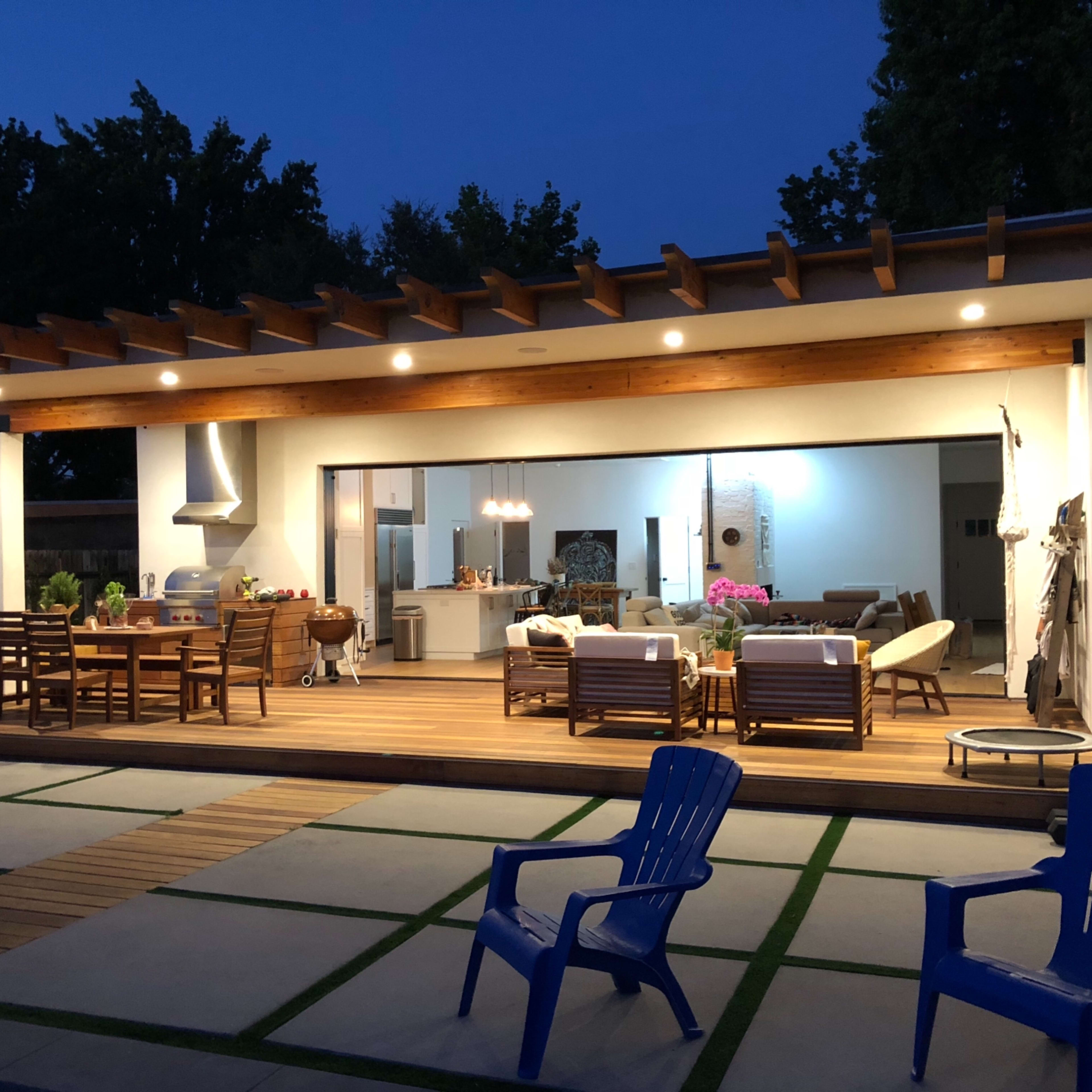 A modern outdoor patio features a wooden deck with dining and lounge areas, surrounded by lush greenery and illuminated by soft lighting at dusk.