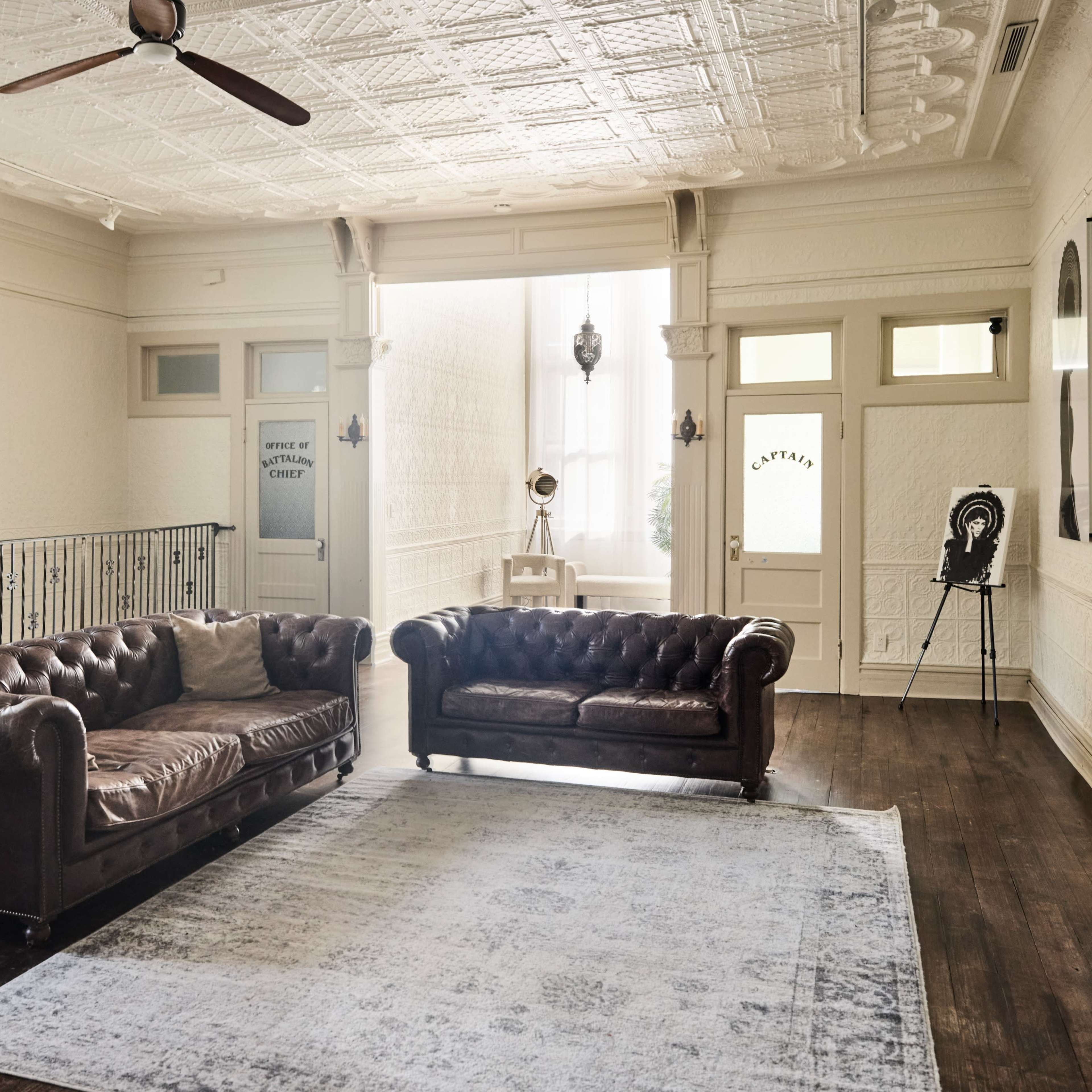 The room features two brown leather sofas and a light-colored area rug on a hardwood floor, with decorative walls and large windows allowing natural light to fill the space.