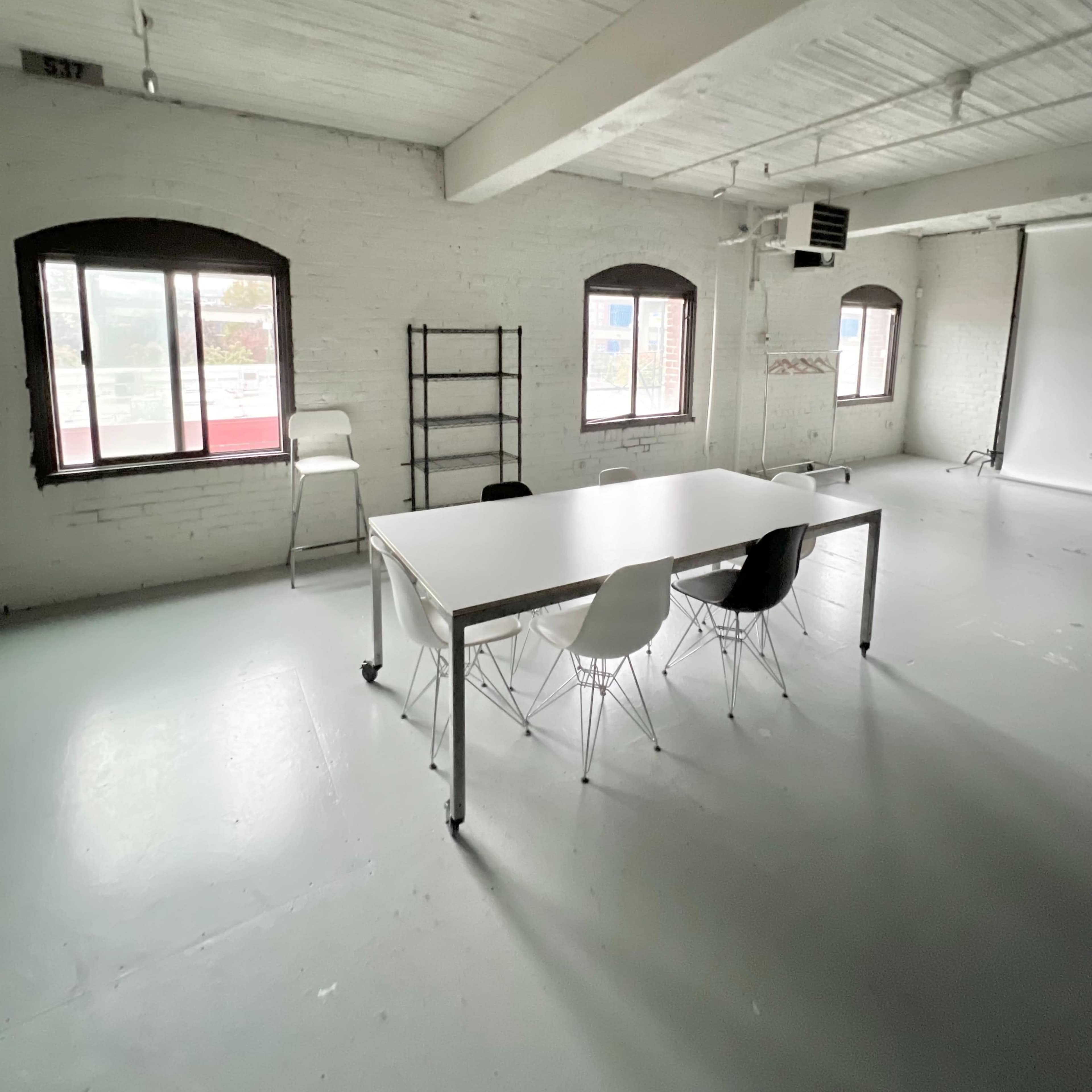 A sparse, well-lit room features a white table and four chairs set against textured walls with large windows.