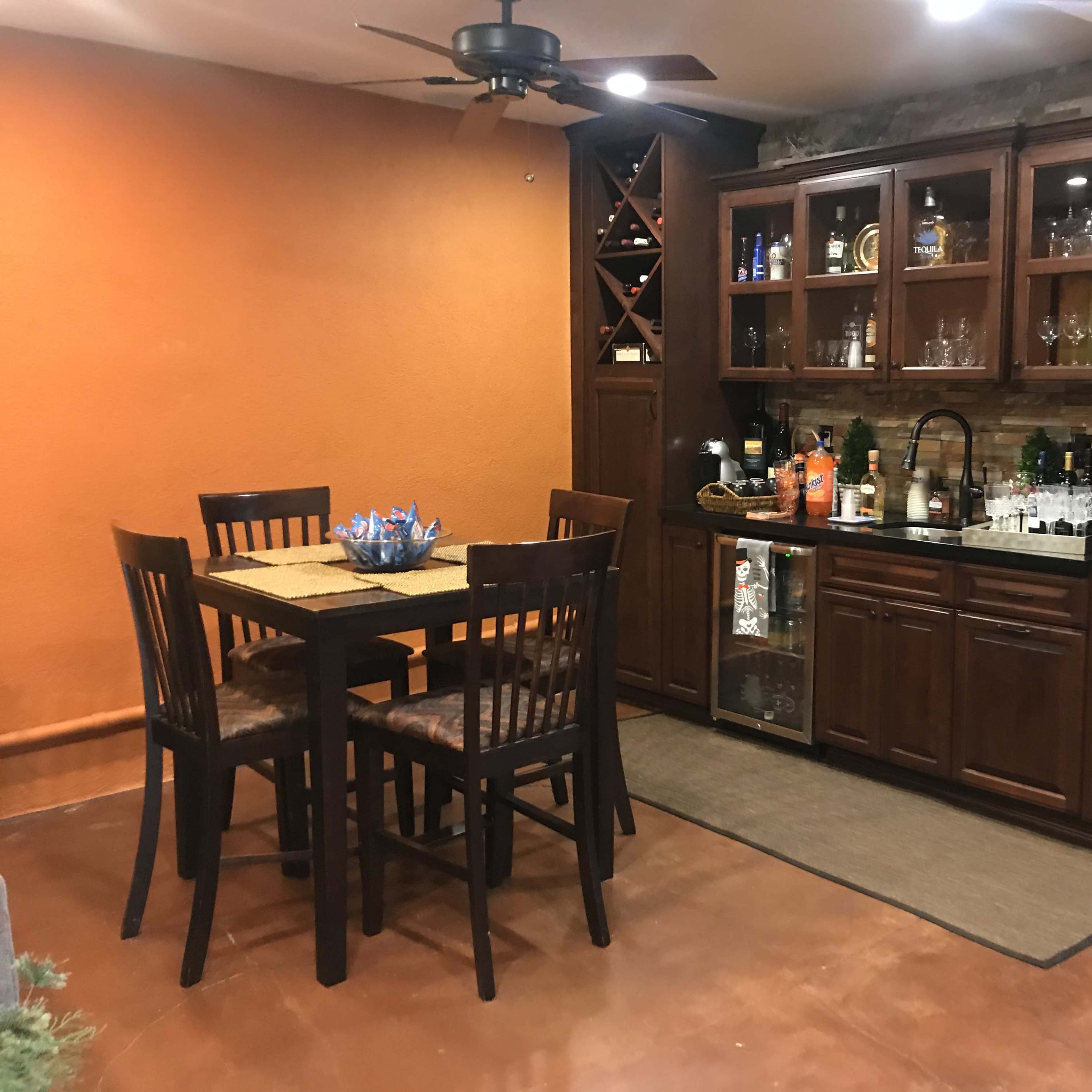 A kitchen area with a wooden dining table and chairs next to a dark wood cabinet featuring a sink, shelves of drinks, and decorative glassware.