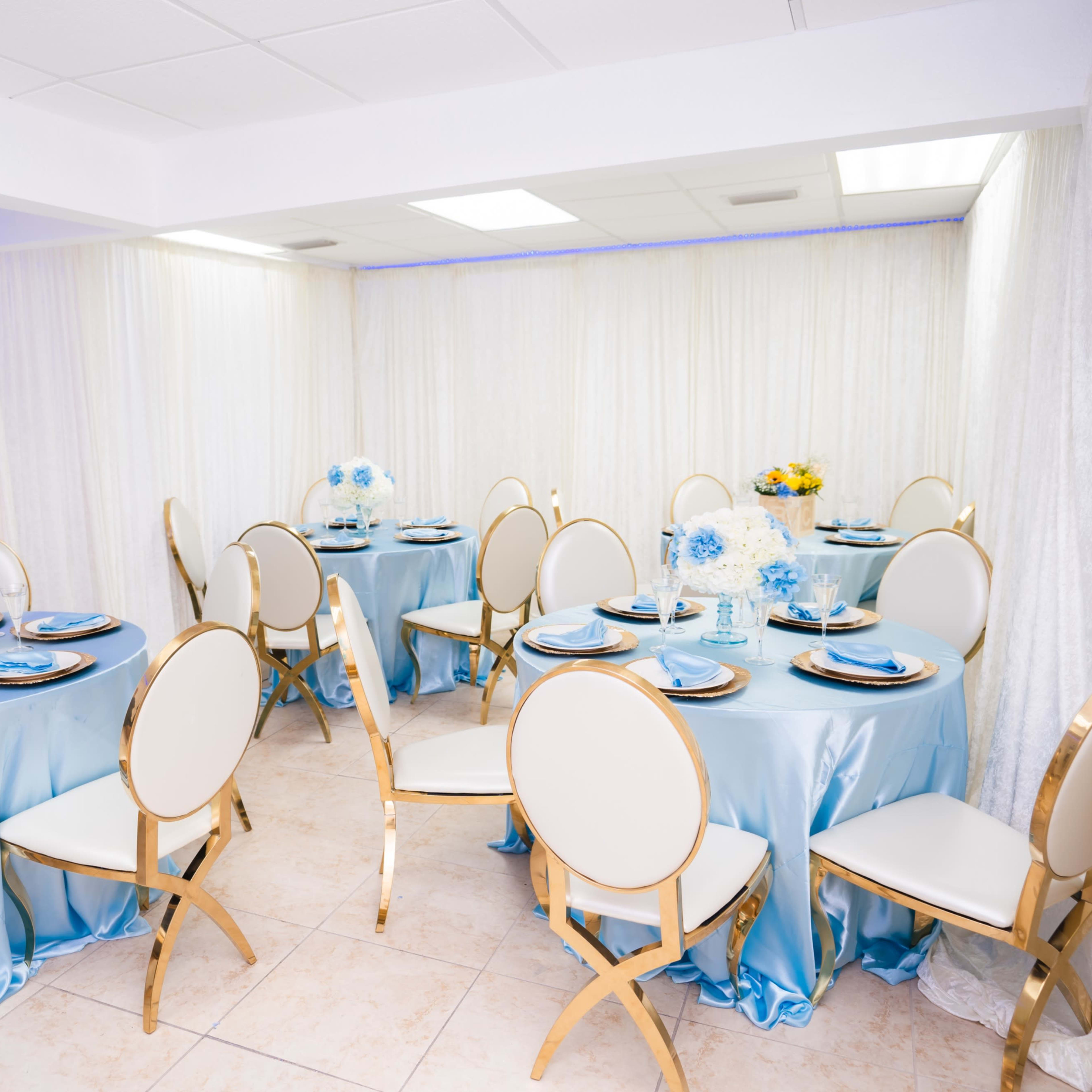 A brightly lit banquet room set up with round tables covered in light blue tablecloths, surrounded by gold-accented chairs.