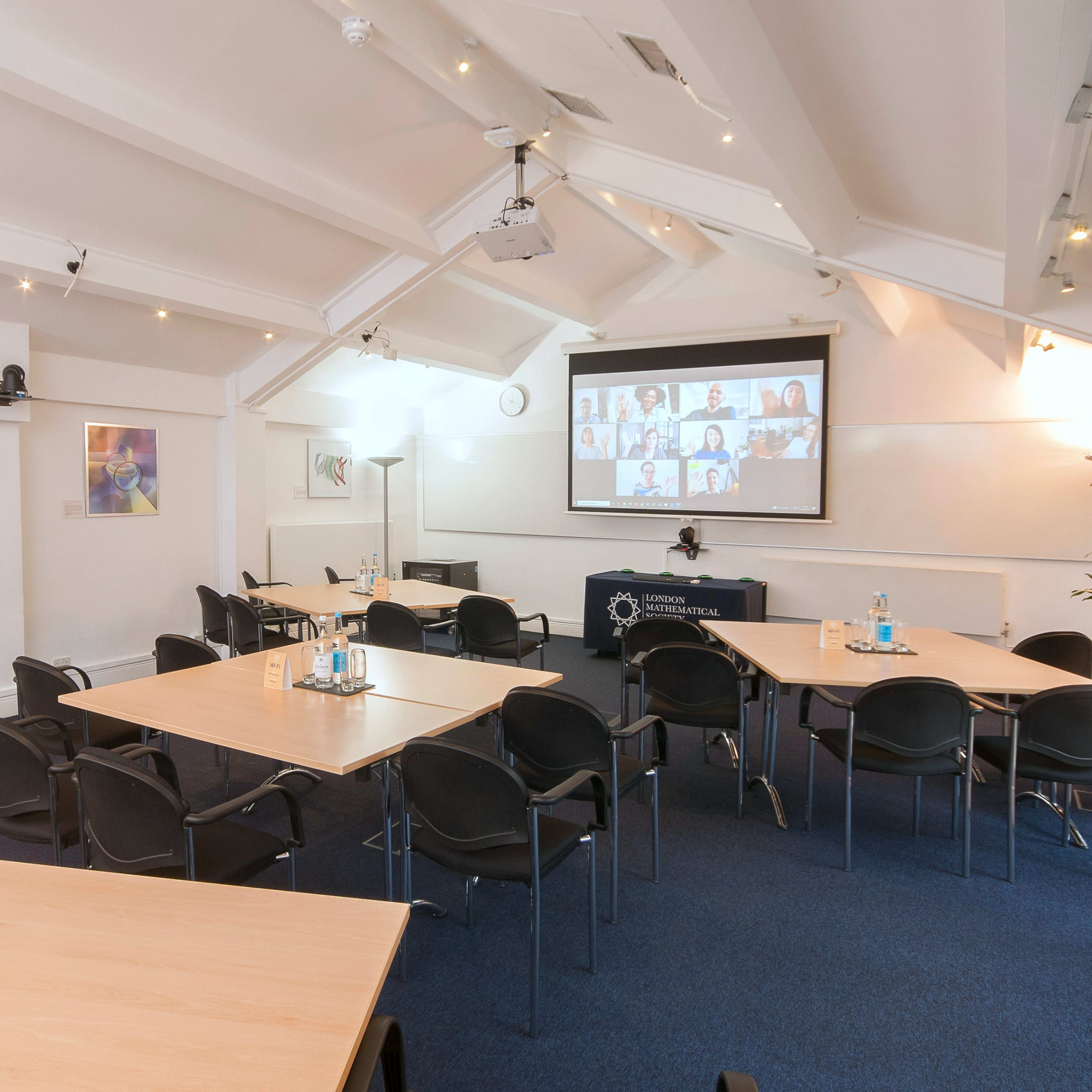 A business meeting room is set up with several tables and chairs, featuring a screen displaying a video call with participants.