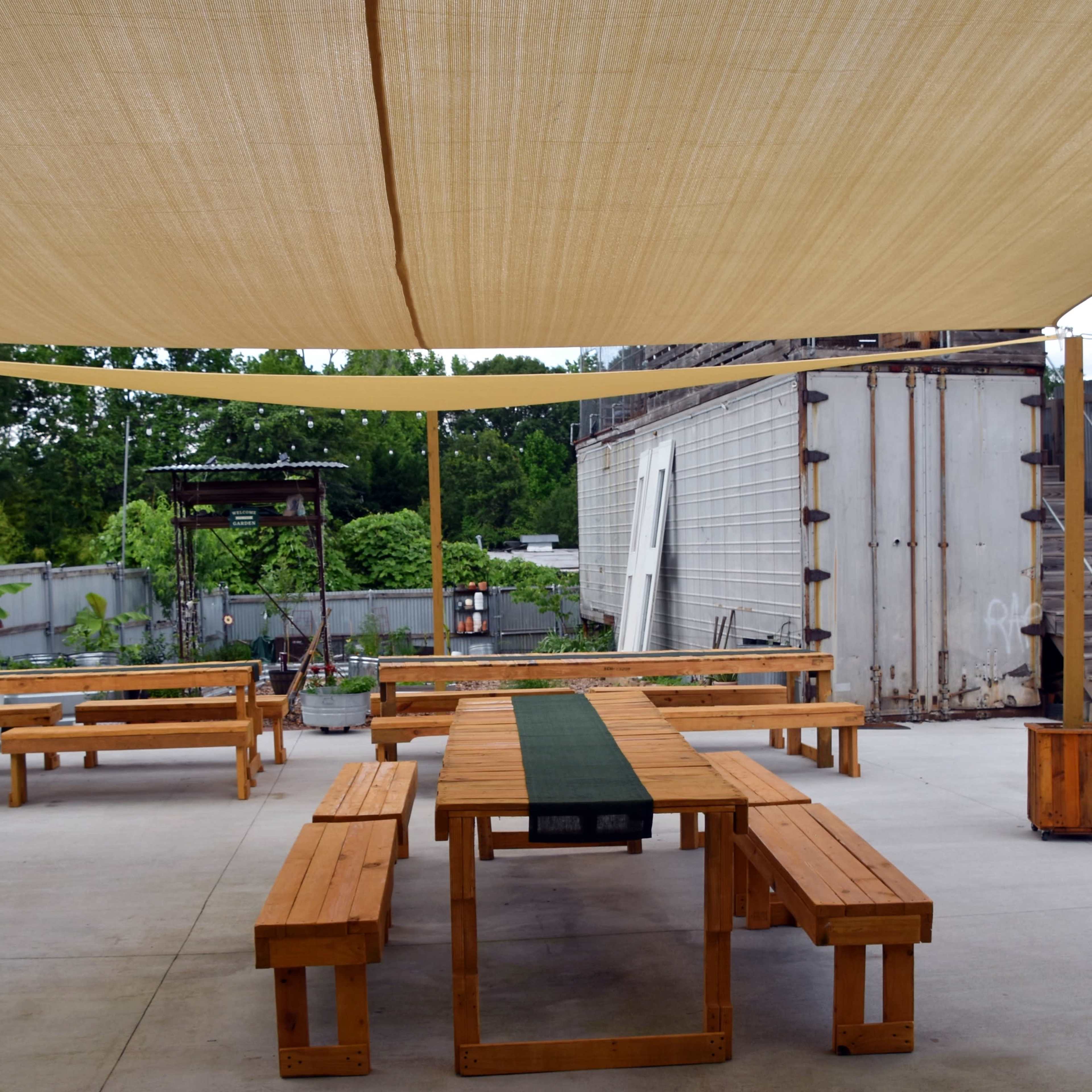An outdoor seating area with wooden tables and benches shaded by a fabric canopy, alongside a storage container and greenery in the background.