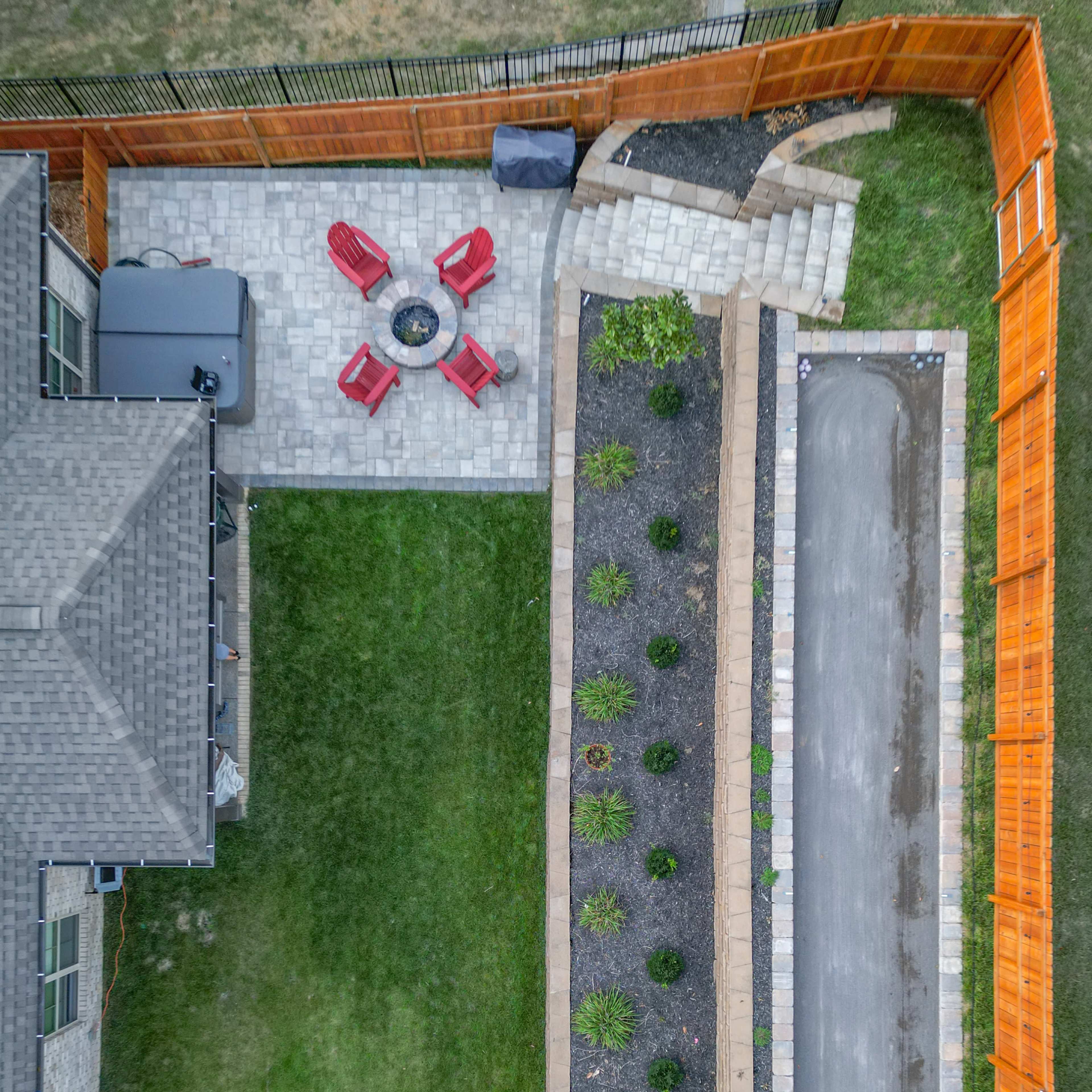 An aerial view shows a backyard with a paved seating area featuring four red chairs around a fire pit, a garden along one side, and a gravel pathway leading to a staircase.