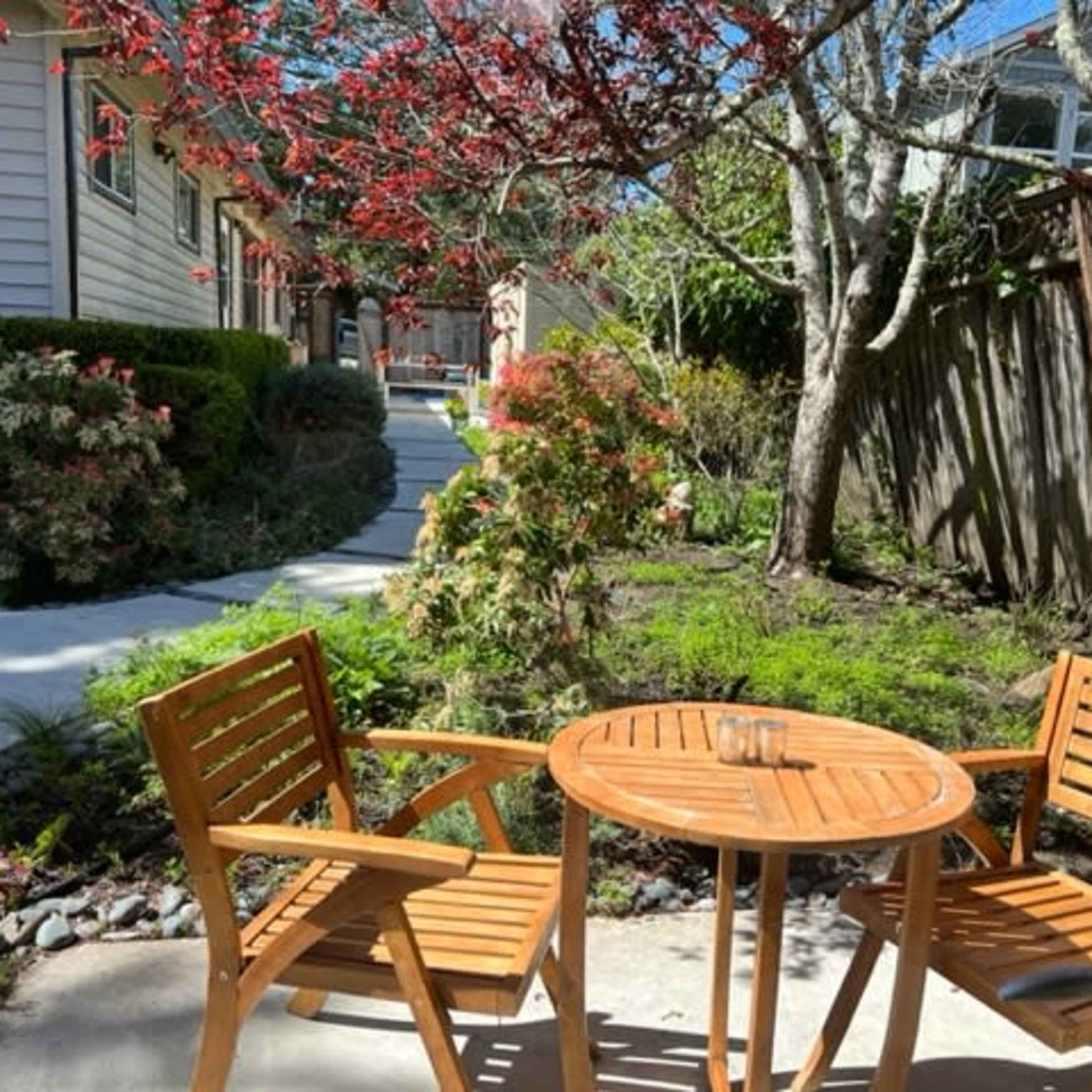 A small patio area with two wooden chairs and a table surrounded by lush greenery and trees.