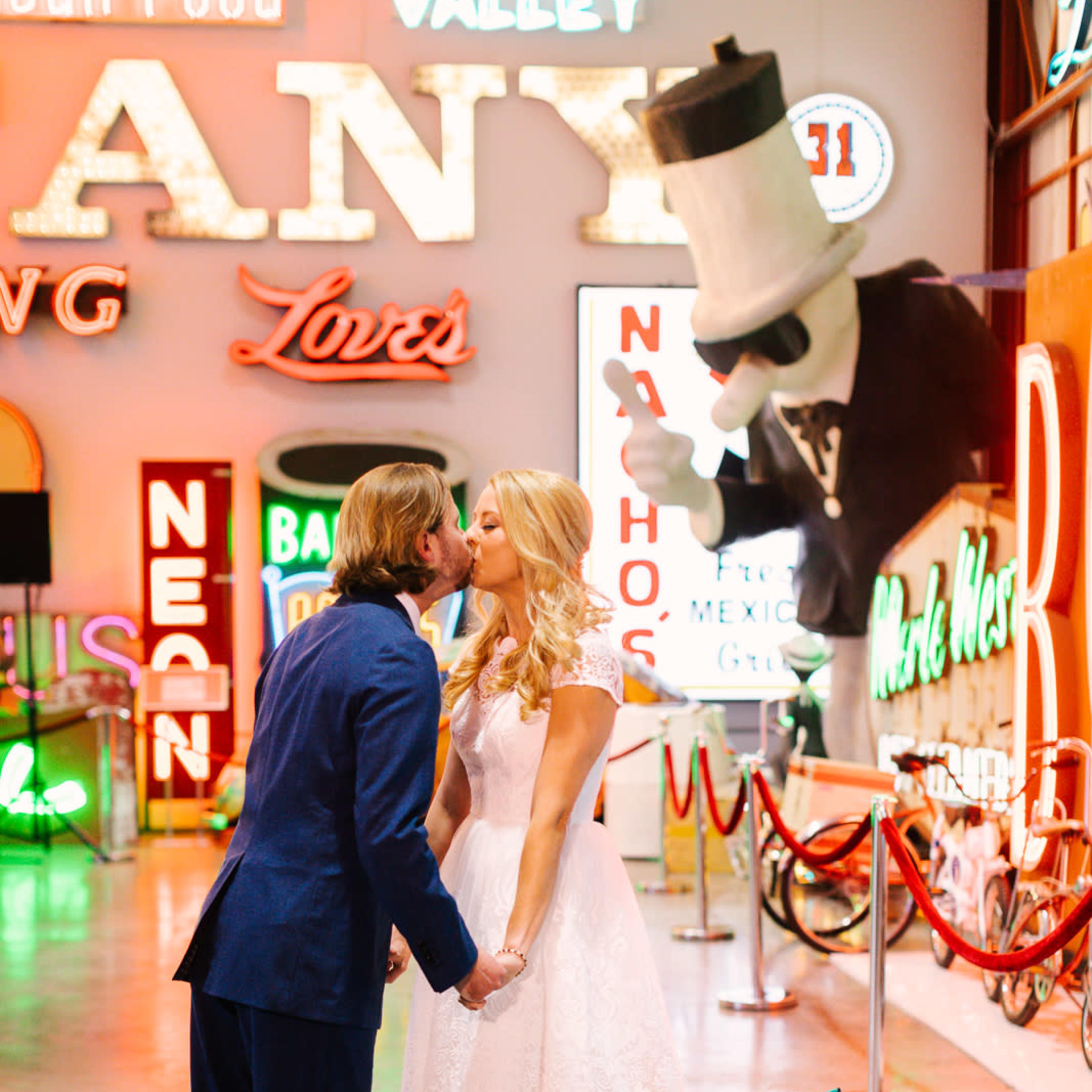 A couple shares a kiss while holding hands in a brightly lit venue adorned with vintage neon signs.