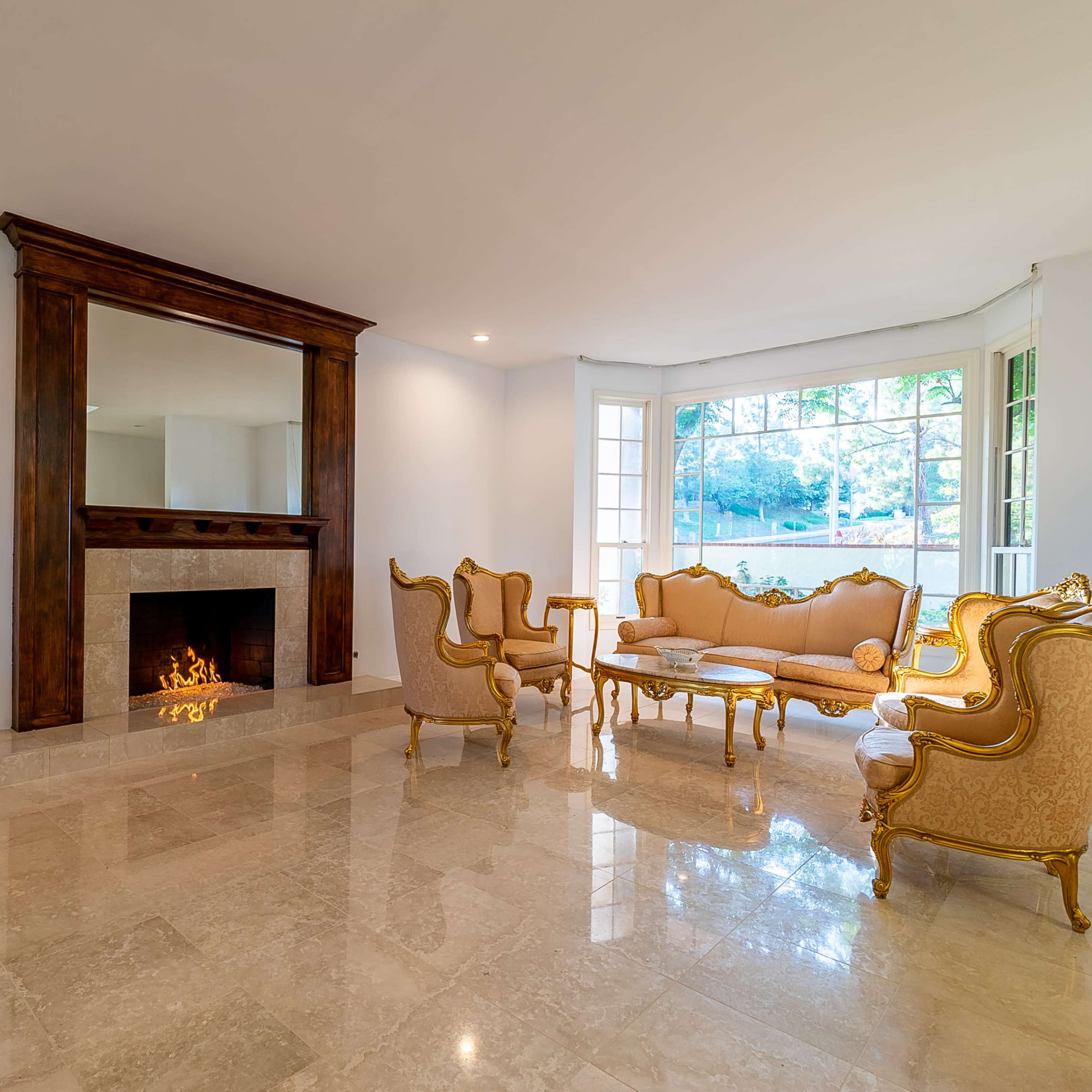 A bright living room with marble flooring, a decorative fireplace, and elegant gold-accented furniture arranged around a central coffee table.