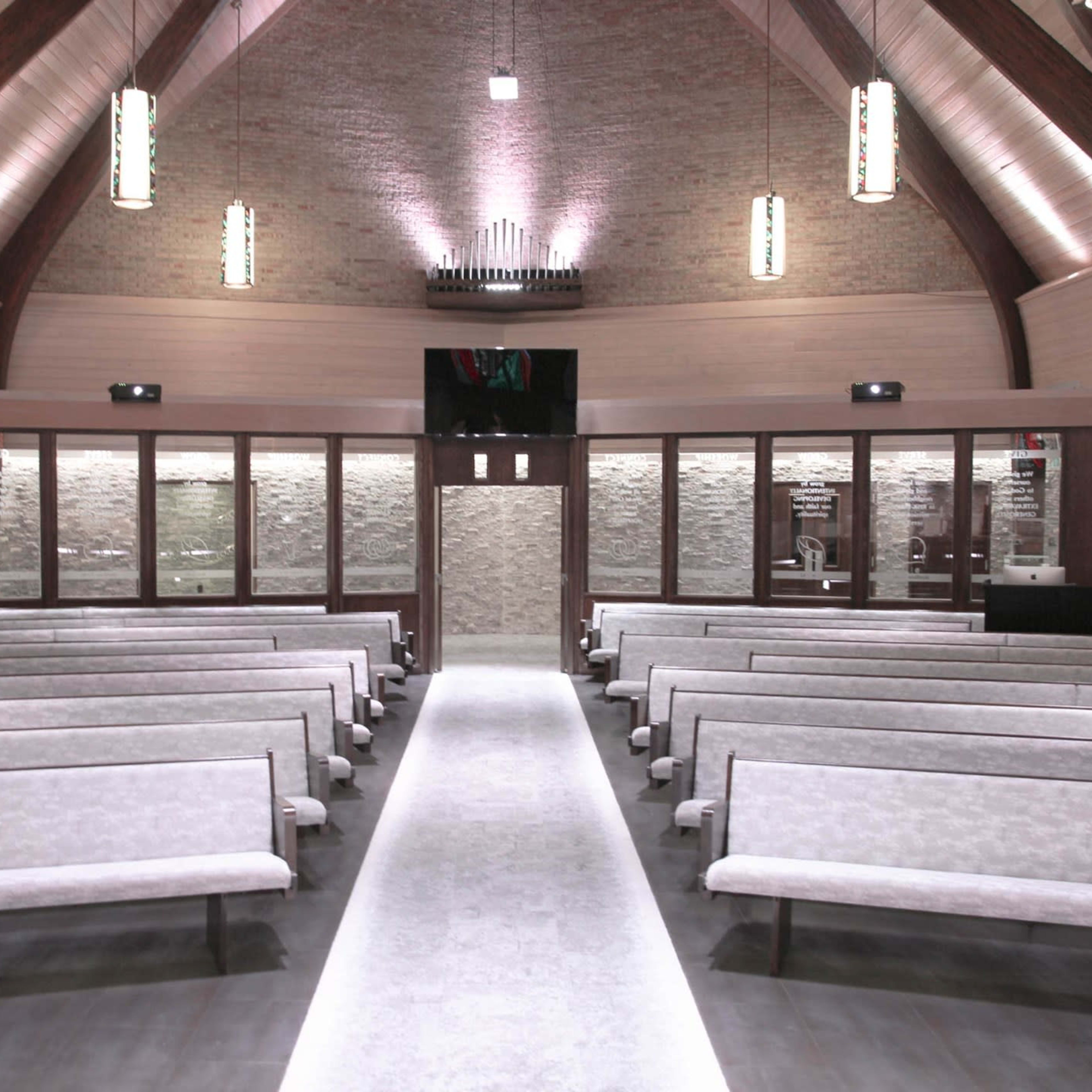 An empty, modern interior of a church with rows of benches and a central aisle leading to large windows and a lighted altar area.