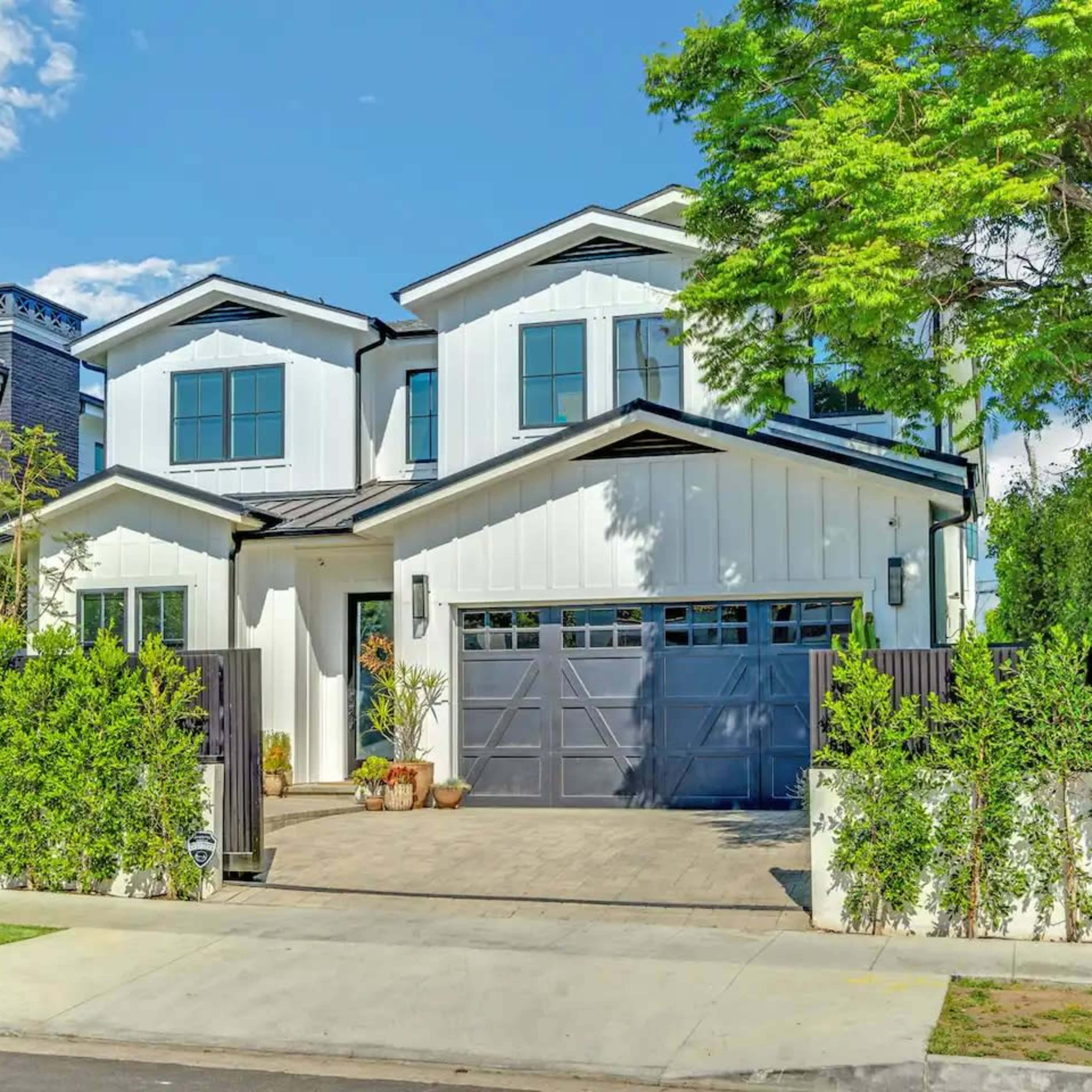 A white two-story house with a black roof and a paved driveway is surrounded by greenery and features a two-car garage.