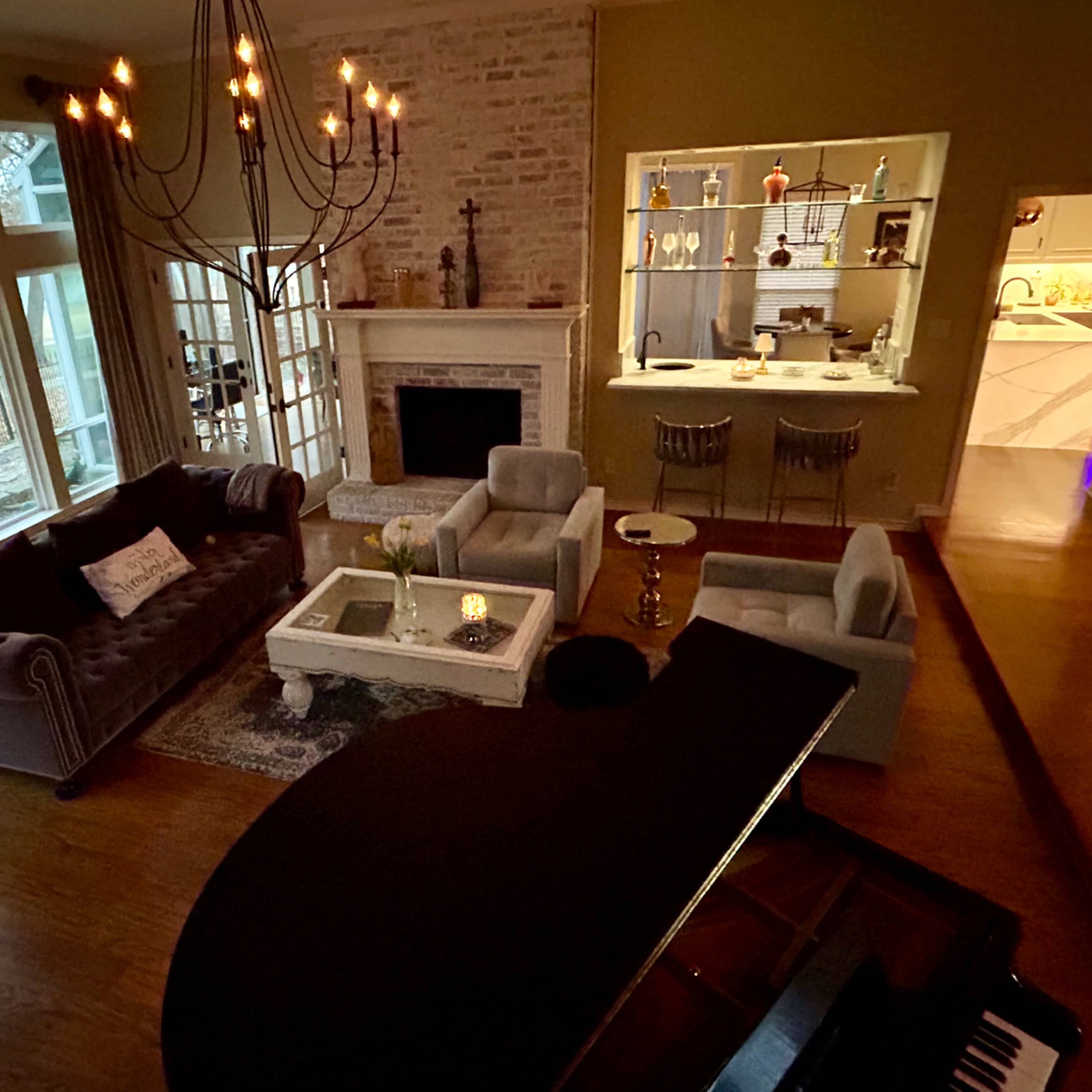 A spacious living room with a grand piano in the foreground, a chandelier above a seating area, and large windows overlooking a scenic view.