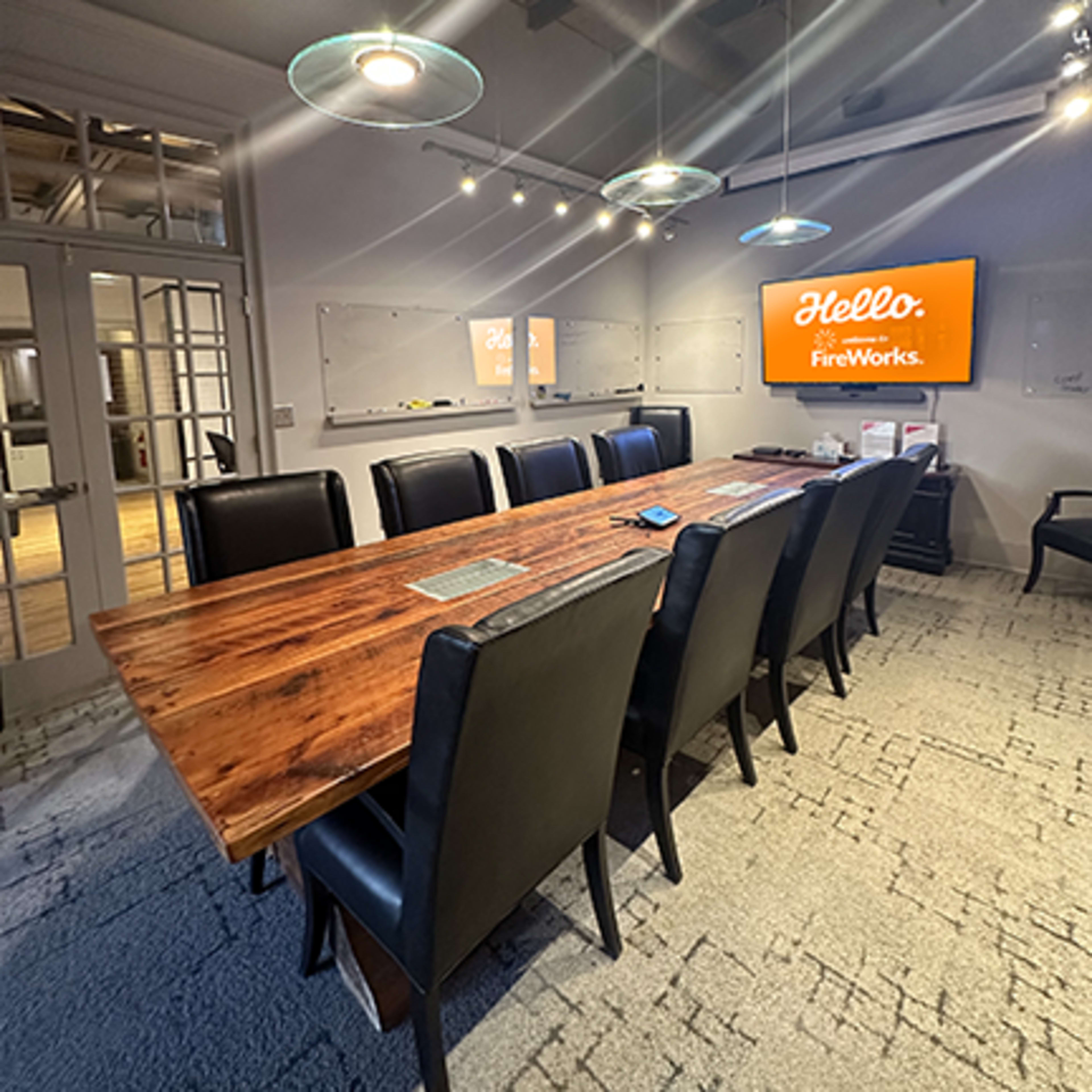 A modern conference room features a large wooden table surrounded by black chairs, with a screen displaying "Hello, FireWorks" on the wall.