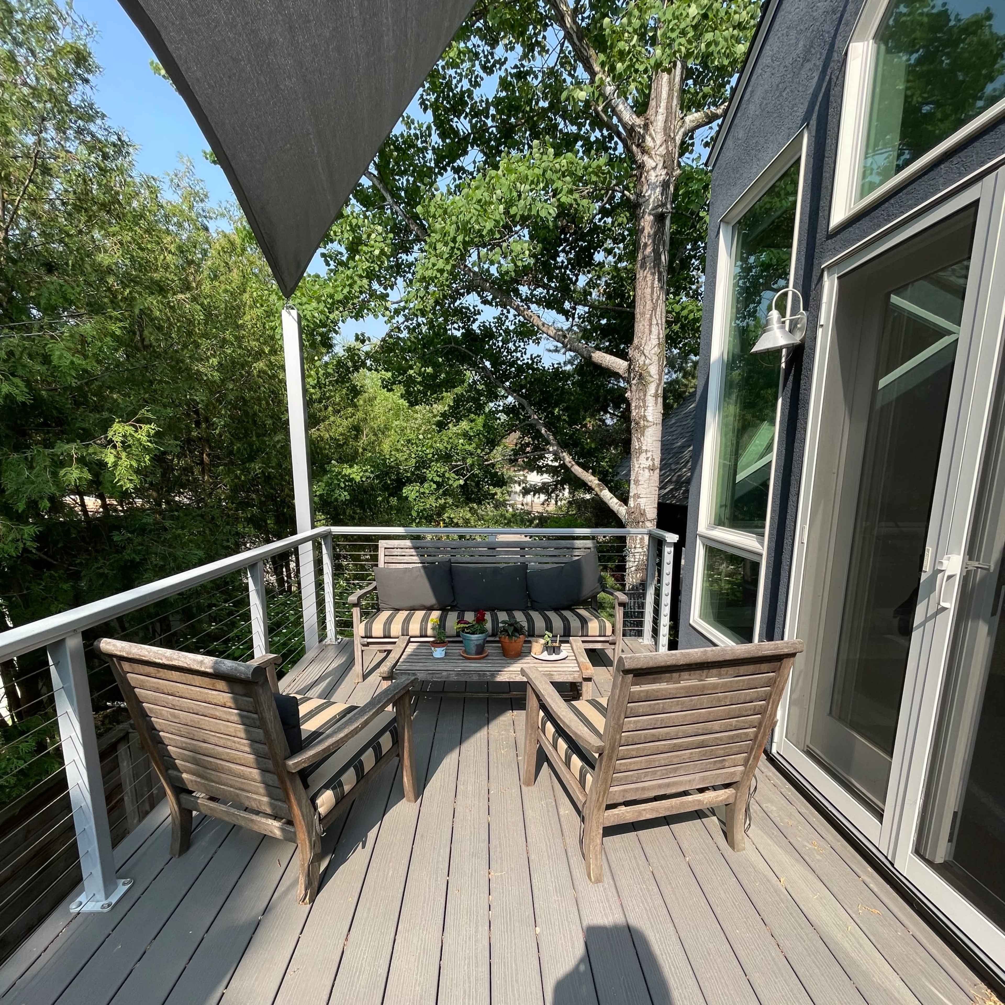 A deck with two wooden chairs and a small table, surrounded by greenery and a sunshade overhead.