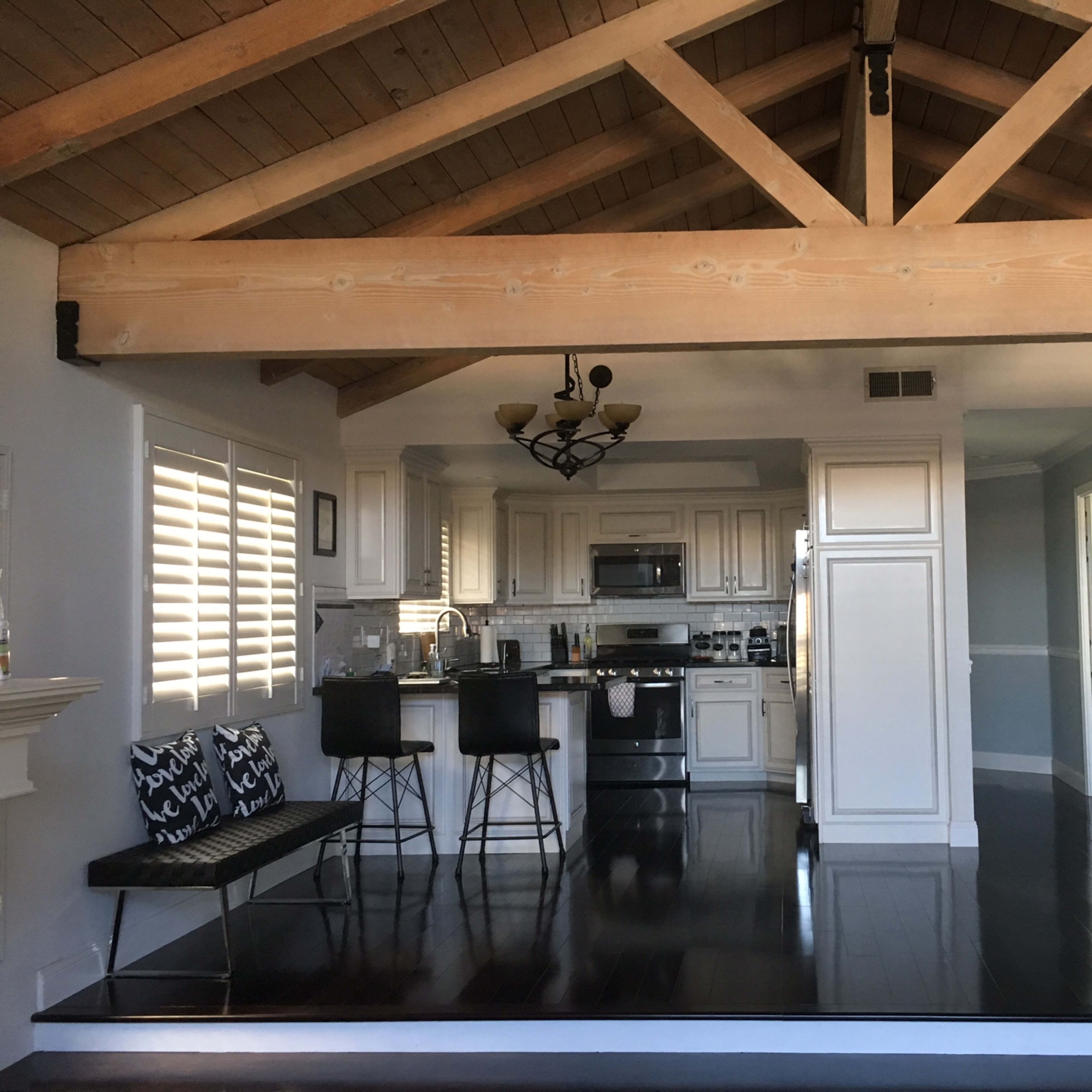 A spacious living area with a kitchen in the background, featuring a high wooden beam ceiling and sleek black flooring.