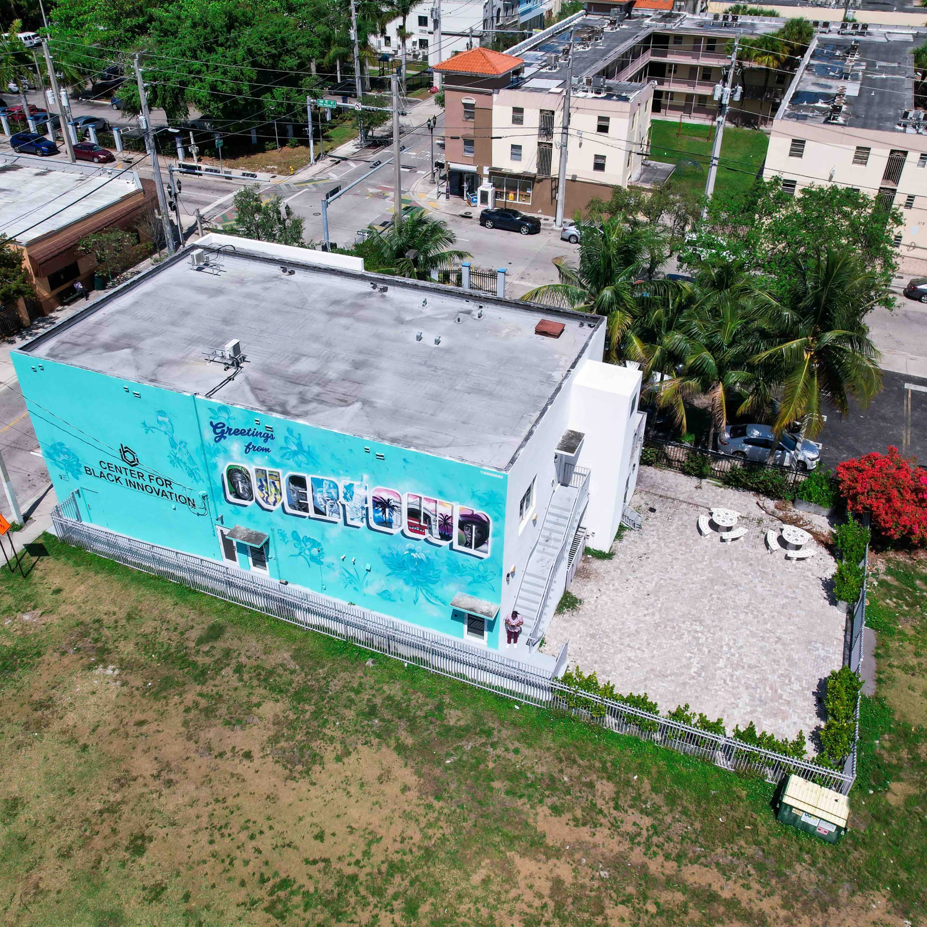 An aerial view shows a building with a colorful mural and a landscaped outdoor area surrounded by residential structures and streets.